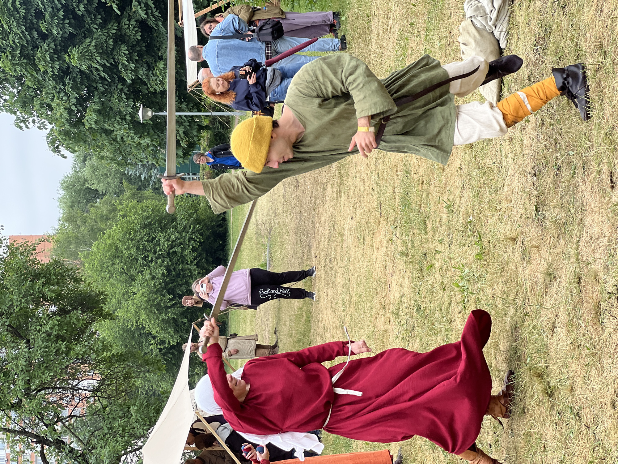 An outdoor reenactment event is underway in Moscow, Russia, possibly at a historical park or festival. Two individuals, one dressed in a long, flowing red medieval-style dress with a white head covering and the other in a green tunic and yellow hat, are engaged in a sword fight demonstration. The person in red is on the left, holding a sword at an angle, and the person in green is on the right, with their sword raised. Both appear to be in mid-action, with the person in red appearing to be in a defensive or offensive stance, and the person in green lunging forward.
Several other people are visible in the background, some watching the demonstration and others appearing to be participants in historical attire. Some are holding cameras and phones, suggesting they are documenting the event. Tents and some scattered historical props or equipment are present, contributing to the reenactment atmosphere. The ground is covered in dry grass, indicating it's likely a warm season. Lush green trees and some buildings are visible in the distance, framing the scene and suggesting an urban or semi-urban park setting. The overall mood is active and participatory, with a focus on historical representation.