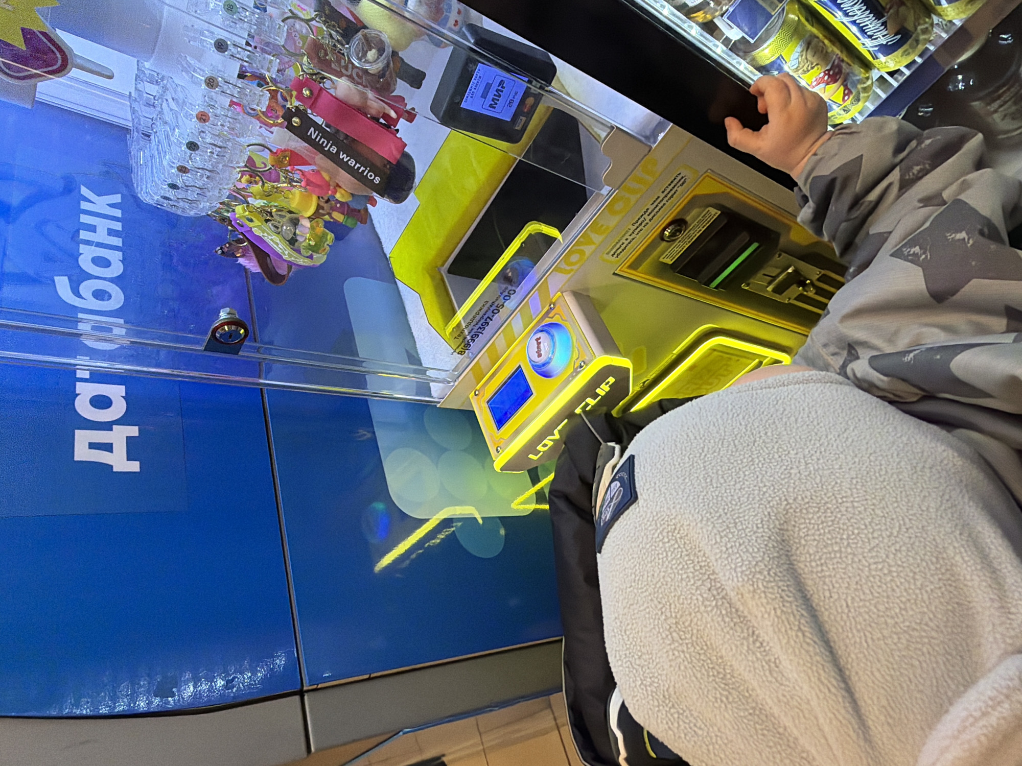 The image shows a child interacting with a prize vending machine in an indoor commercial setting, likely in Votkinsk, Russia. The child, seen from an overhead angle, wears a grey camouflage jacket and a light grey fleece top, with their hand near the machine's interface, suggesting they are playing or about to play the game.
The vending machine is brightly lit, featuring a transparent case filled with various keychains as prizes, including one clearly labeled "Ninja warrios." The machine's control panel has a prominent blue "Start" button and a display showing "600" and the brand "LOVE CLIP."
Payment options include a terminal inside the machine displaying "МИР" (MIR), Russia's national payment system, and the number "20." A separate panel on the machine provides "Техподдержка" (Technical Support) information, including "Telegram/WhatsApp" and the phone number 8(999)397-05-00.
To the left of the machine, a blue panel displays the Russian text "Да!Банк" (Da!Bank). In the background, blurred items suggest other merchandise, possibly canned drinks, indicative of a shop, arcade, or convenience store environment with artificial lighting. The scene captures a moment of playful engagement with the arcade game.