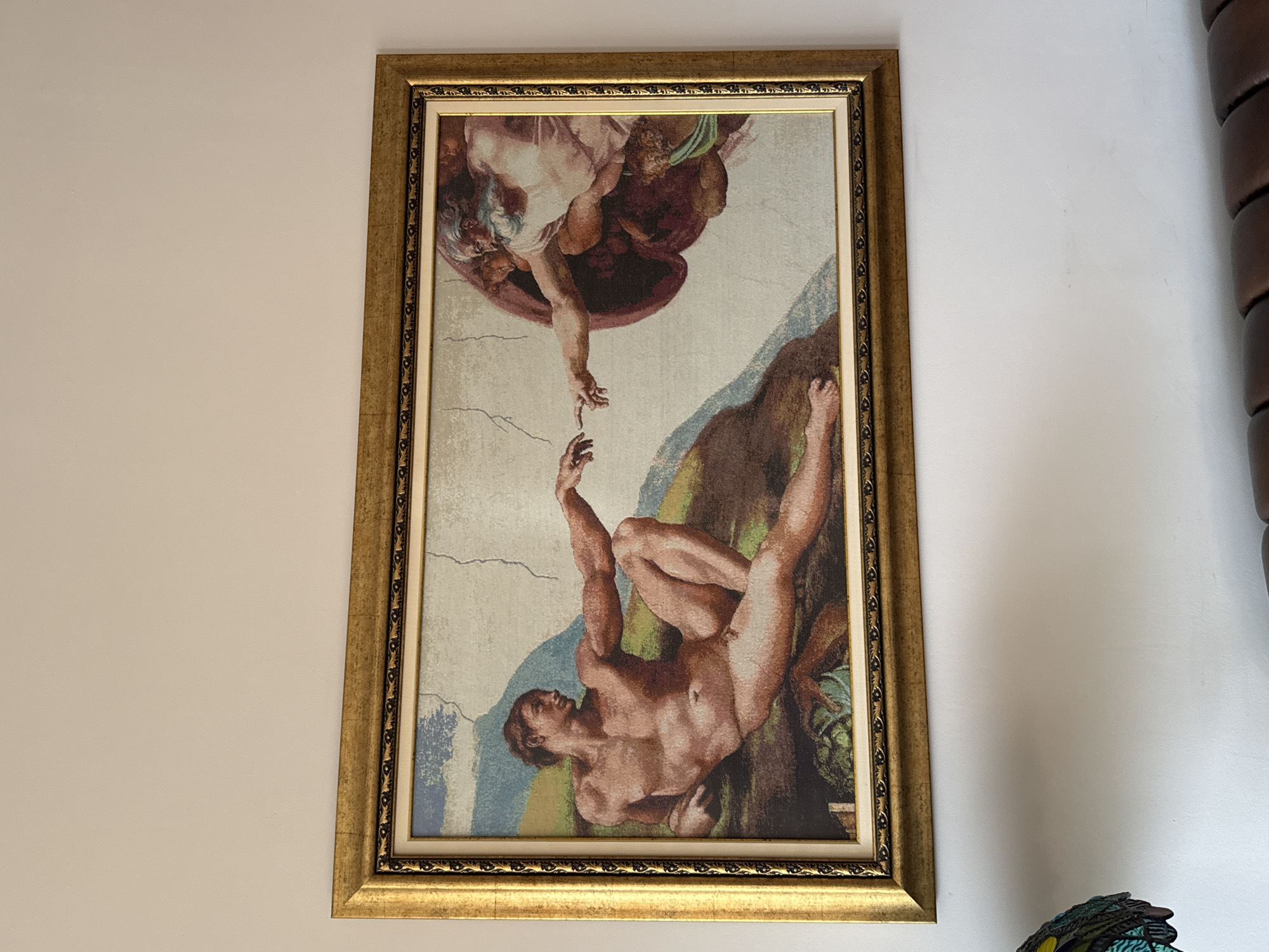 A framed tapestry of Michelangelo's "The Creation of Adam" hangs on a white wall. The tapestry is rectangular and depicts the famous scene of God reaching out to Adam. The frame is gold with an ornate design. The tapestry itself appears to be a woven textile, with some visible texture. To the right of the frame is a dark brown, vertically-oriented object. The tapestry and frame are the primary focal point. No information suggests that the image was taken in Plovdiv, Bulgaria.