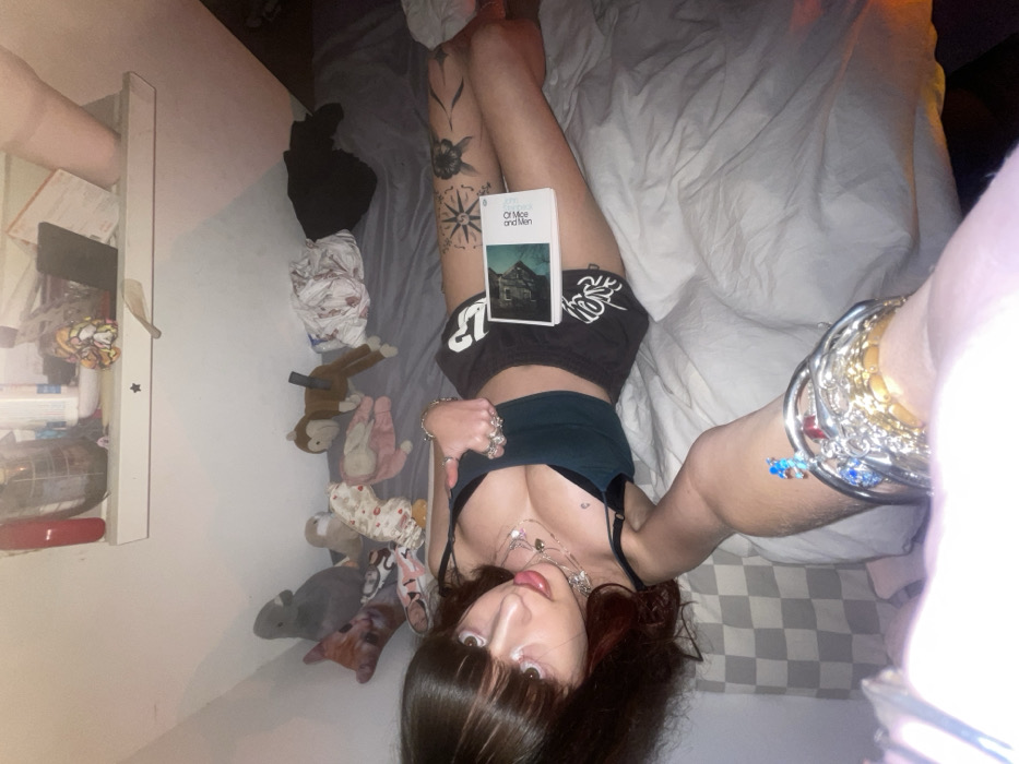 A woman reclines on a bed with white sheets. She holds a book titled "Of Mice and Men" on her lap. She is wearing a green top and dark shorts. She has multiple tattoos on her legs. Her hair is dark with bangs. She has multiple bracelets on her wrist. There are stuffed animals and other objects visible near the bed. A white shelf is attached to the wall.