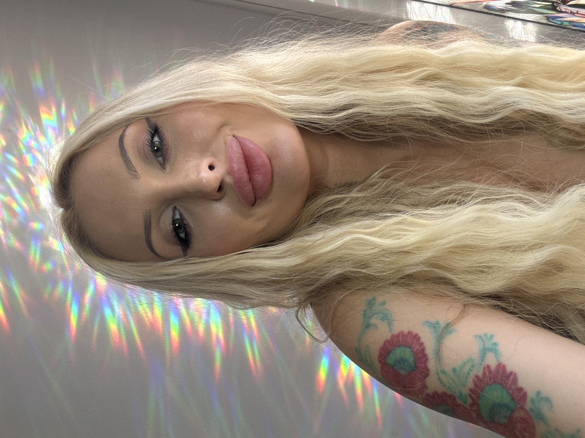 An overhead, medium shot shows a blonde woman looking up and to her right, towards the camera, in an indoor setting. The woman has long, wavy blonde hair that falls over her shoulders. She has bold makeup, including dark eyeliner and noticeable lip filler. Her left arm, visible on the left side of the frame, has a tattoo of stylized flowers in shades of pink and teal. The background is a light-colored wall on which colorful light patterns are cast, resembling streaks of rainbow light. The environment appears to be a room, possibly a home, as indicated by the lighting and general atmosphere. No specific location in a city is discernible.