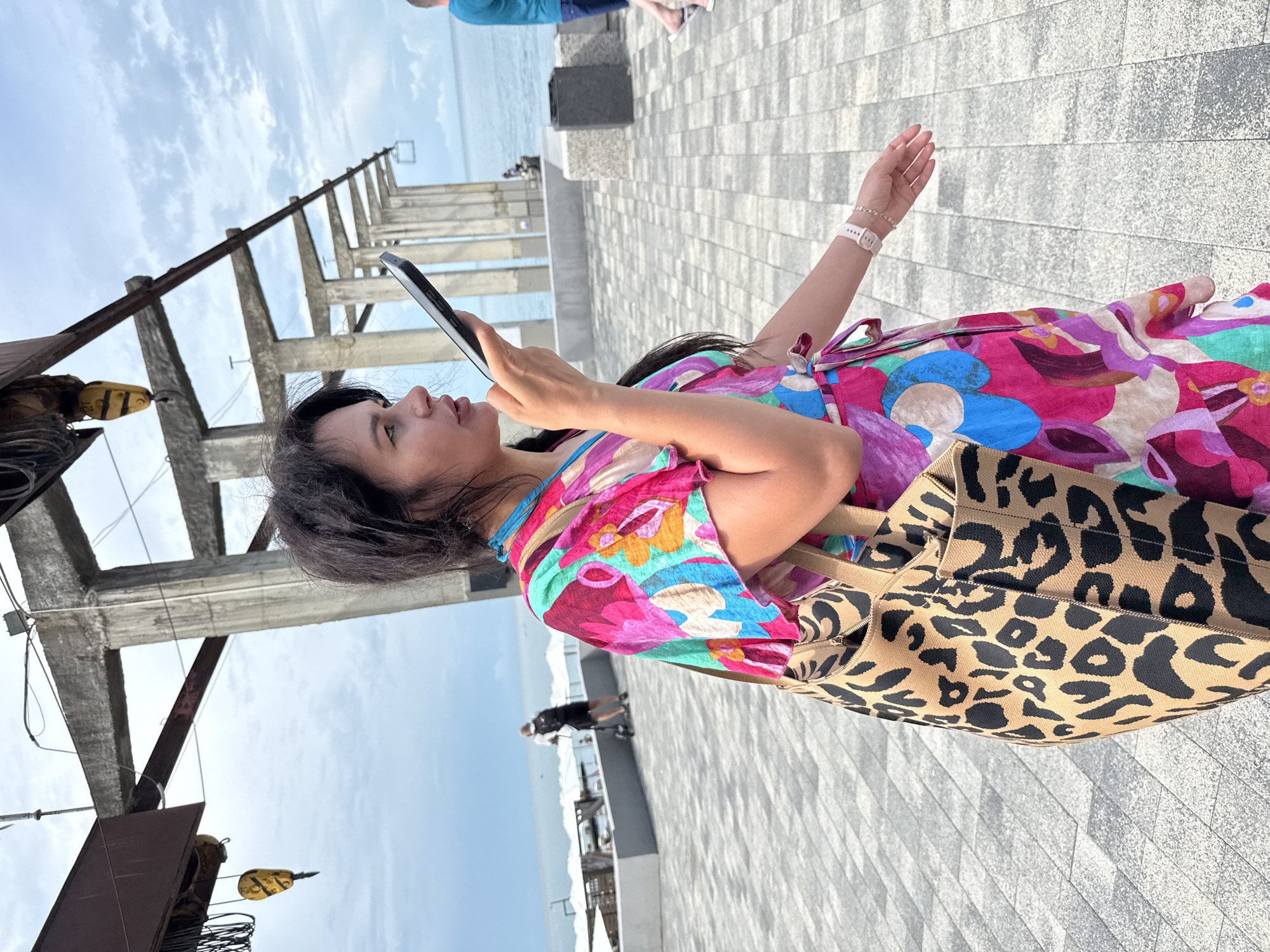 A woman with long, dark hair is walking on a paved walkway near the sea, holding a smartphone to her ear as if speaking into it. She is wearing a vibrant, multi-colored dress with a floral pattern and carrying a large leopard-print tote bag over her left shoulder. In the background, a man on a scooter is visible, and further out, the sea stretches to the horizon under a cloudy sky. Concrete structures, likely part of a pier or an industrial area, are present in the background. The scene appears to be outdoors in a public space with a view of the ocean.