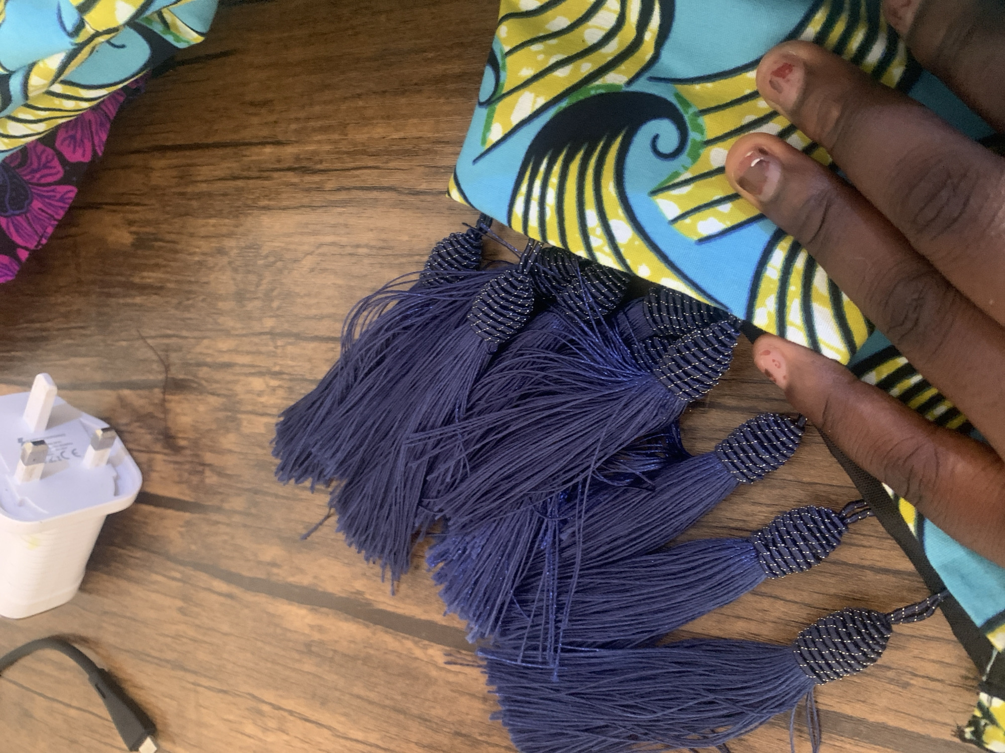 A close-up shot shows a collection of deep blue tassels with beaded tops arranged on a wooden surface, possibly a table or floor. To the right, a hand with dark skin and chipped nail polish is seen resting on a piece of fabric with a vibrant, abstract pattern of yellow, blue, and black swirls. In the upper left corner, a white power adapter with three metal prongs and a black USB cable are visible. The scene appears to be an indoor setting, likely in Jauro Yinu, Nigeria, given the context provided.