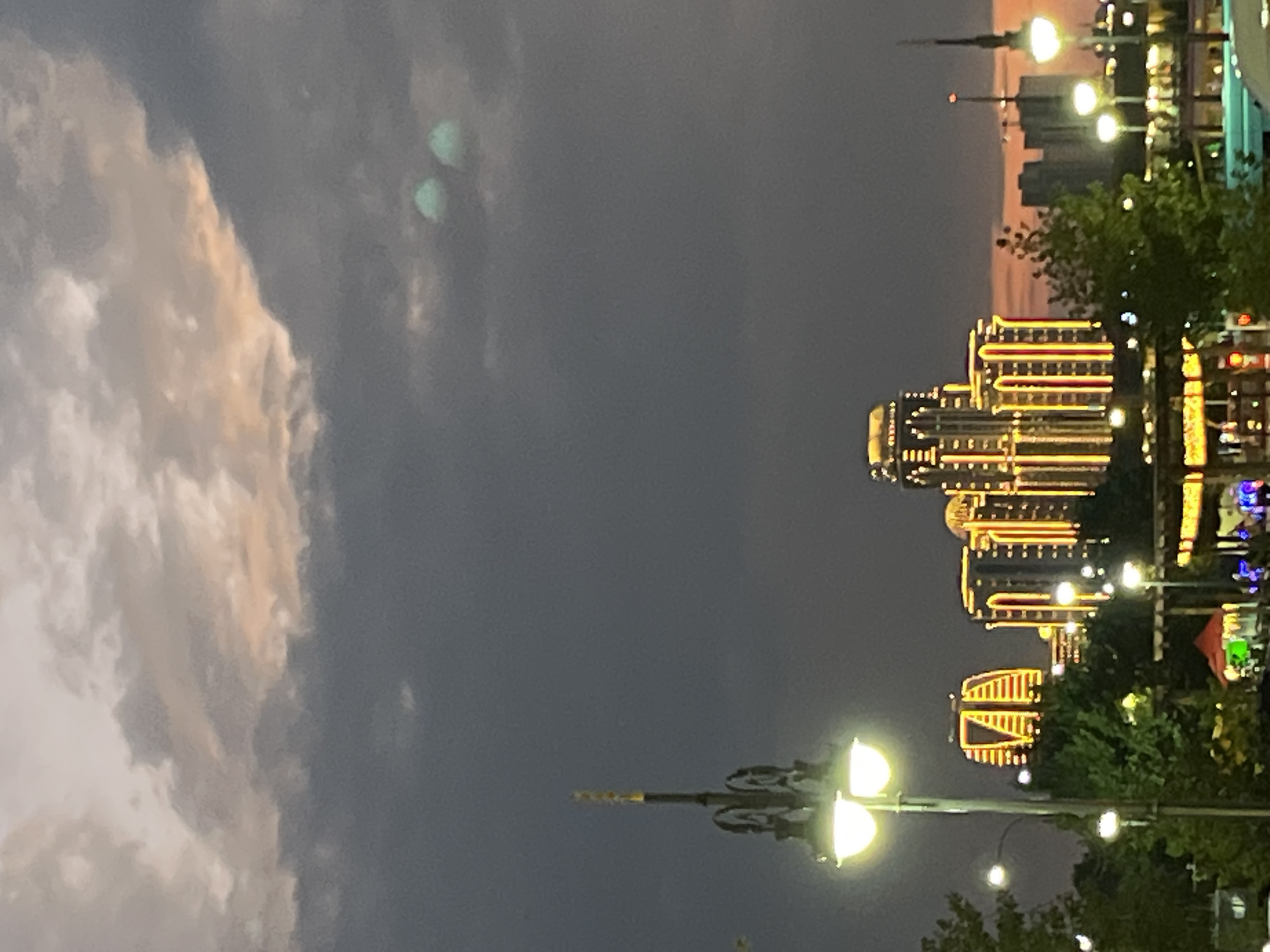 In Grozny, Russia, at dusk, an array of illuminated skyscrapers stands against a dramatic, cloudy sky. The buildings are adorned with vertical lines of bright yellow and red lights, creating a striking contrast with the darkening atmosphere. A vintage-style lamppost with two glowing orbs stands in the foreground, casting a warm light. Trees and other smaller structures are visible between the foreground and the skyscrapers, suggesting a city park or promenade. The sky is a mix of deep grays, purples, and a faint peachy hue near the horizon, with two blurry, pale green orbs hovering in the upper right portion of the frame. The overall scene captures the vibrant nighttime cityscape of Grozny.