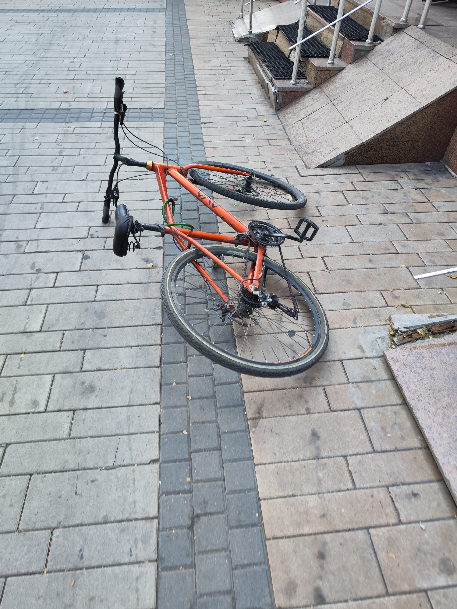 An orange bicycle is lying on its side on a tiled pedestrian walkway. The bicycle appears to be intact and shows no obvious signs of damage. The walkway is paved with rectangular grey and dark grey tiles, arranged in a pattern. To the right of the walkway, there are stone steps leading up to a building. The scene is well-lit, suggesting it is daytime. The image does not contain any people or animals. The overall impression is of an unattended bicycle in a public urban space. This scene could be in Astana, Kazakhstan.