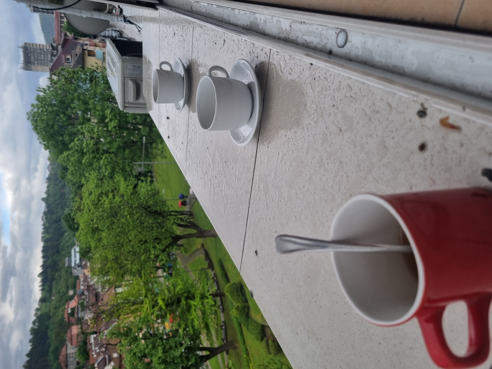 The image shows a view from a balcony in Gabrovo, Bulgaria. Three coffee cups sit on a wet surface, likely an exterior windowsill or ledge. Two are white with matching saucers, and one is a larger red mug containing a spoon. Beyond the ledge, a cityscape unfolds, featuring buildings, trees, and grassy areas. The sky is overcast.