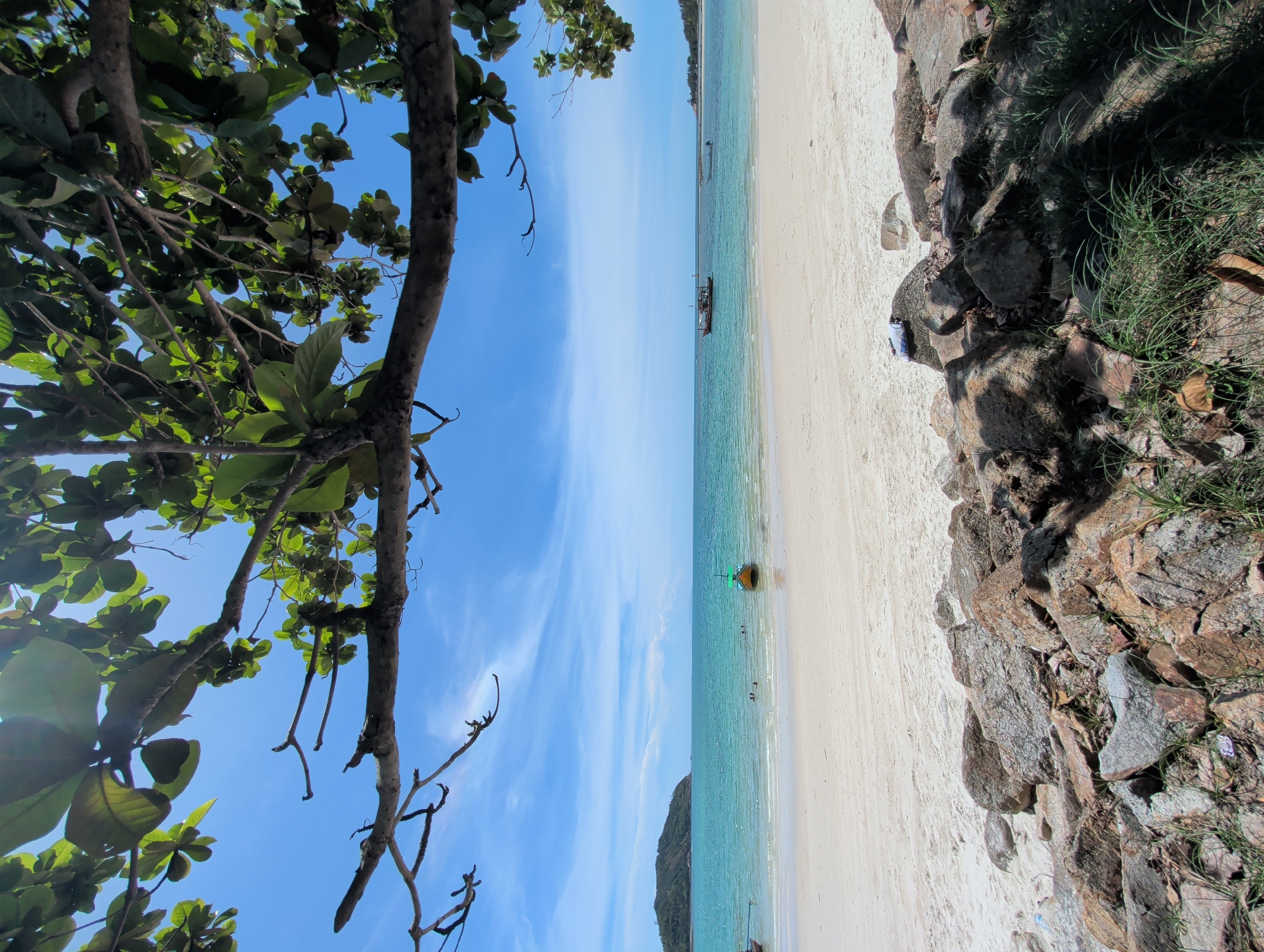 The image shows a beach in ตำบลเกาะพะงัน, Thailand. The scene is framed by trees and rocks. A sandy beach meets clear turquoise water under a blue sky. Several boats are visible offshore, and a few people are in the water. The lighting is bright, suggesting it is daytime.