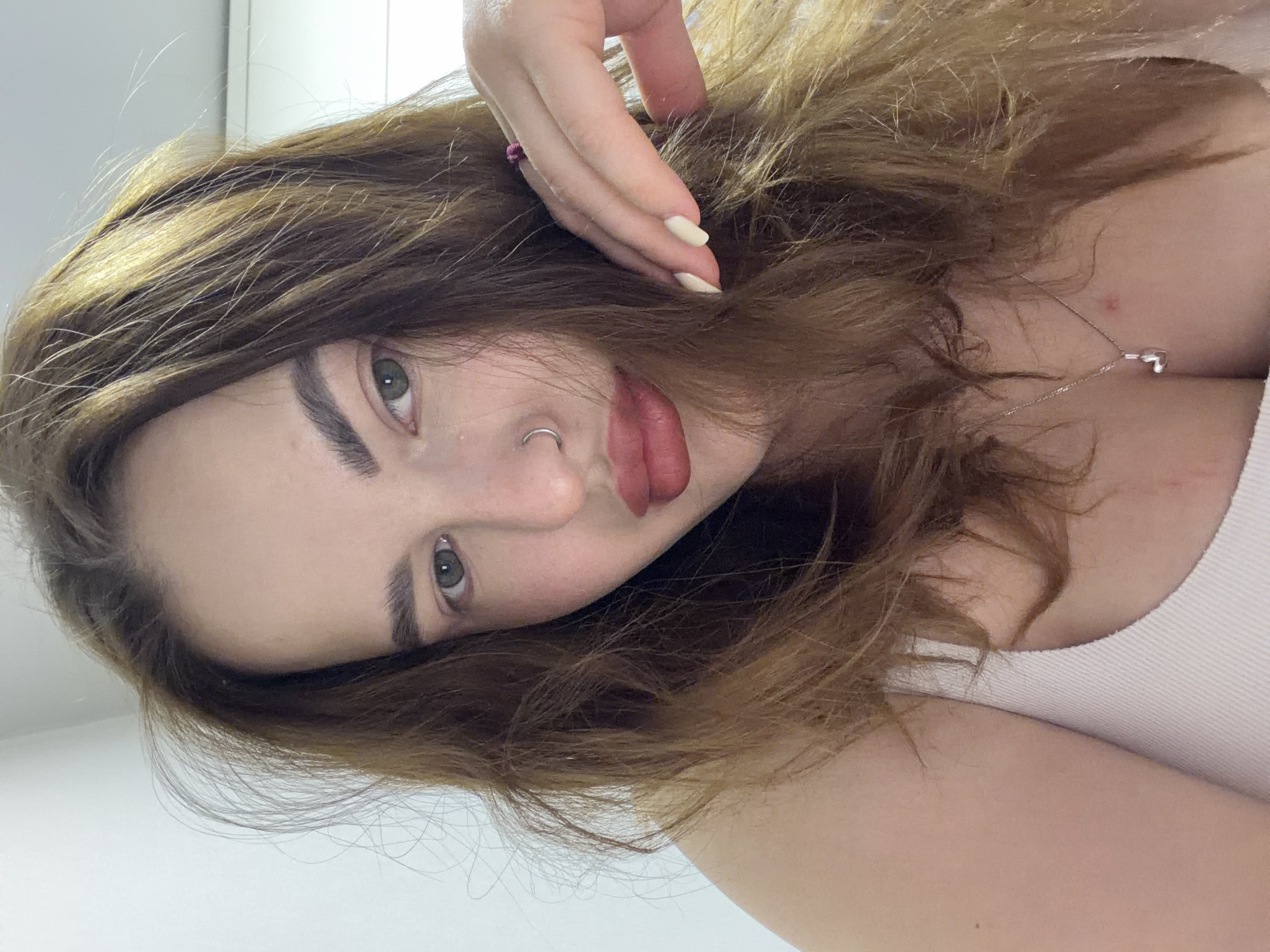 A close-up portrait shows a young woman looking directly at the camera. She has long, wavy brown hair with lighter highlights, which is somewhat disheveled and frames her face. Her eyes are a striking green or hazel, and she has well-defined eyebrows. Her lips are full and painted with a muted red or berry-toned lipstick, slightly parted. She wears a silver hoop nose ring on her left nostril.
Her right hand is visible, with manicured, light-colored nails, resting casually on the top of her head near her temple, gently touching her hair. A small pink hair tie or ring is on one of her fingers. Around her neck, a thin silver chain necklace with a small heart-shaped pendant is visible, partially obscured by her hair and the neckline of her clothing.
She is wearing a light-colored, possibly white or light pink, ribbed tank top or camisole. The background is a plain, light-colored wall or surface, suggesting an indoor setting without any specific distinguishing features. The lighting appears soft and natural, indicating it is likely daytime. Her expression is neutral to slightly contemplative, meeting the camera's gaze. No text is visible in the image. This scene is contextualized as being in Krasnodar, Russia.