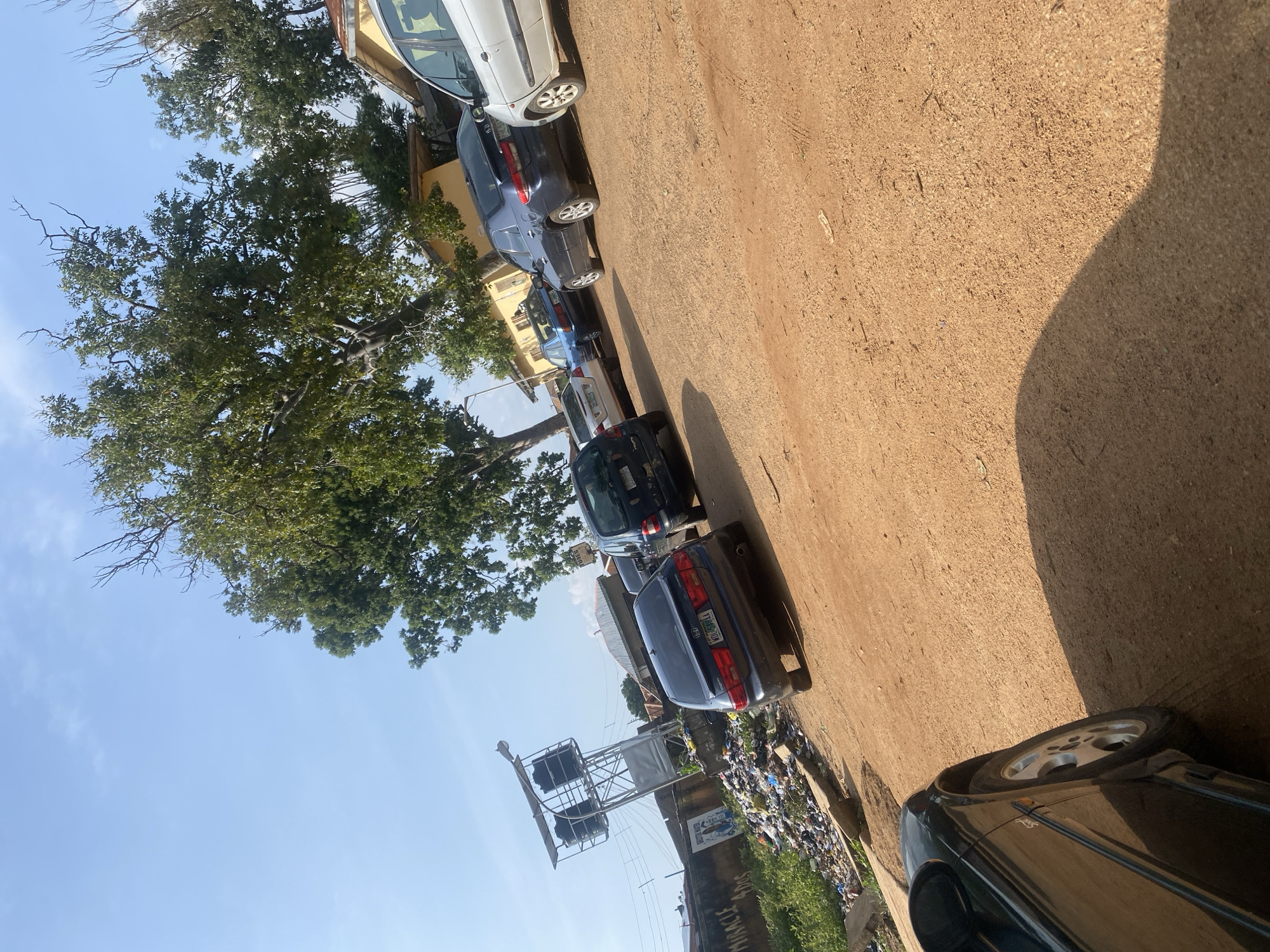 The image depicts an outdoor scene in Rafin Par, Nigeria, characterized by a dirt road lined with parked vehicles and vegetation. In the foreground, a black car is partially visible, casting a shadow on the dusty ground. Behind it, several other cars are parked in a row, including a dark blue sedan and a silver minivan. A few more vehicles are further down the road. To the left of the road, there is a pile of garbage, with a water tower structure visible behind it. A weathered concrete wall with some graffiti runs along the side of the garbage pile. The background features several buildings with yellow walls and corrugated metal roofs, interspersed with trees providing shade. The sky is clear and blue, indicating a sunny day. The overall environment suggests a residential or semi-commercial area with visible signs of waste accumulation.