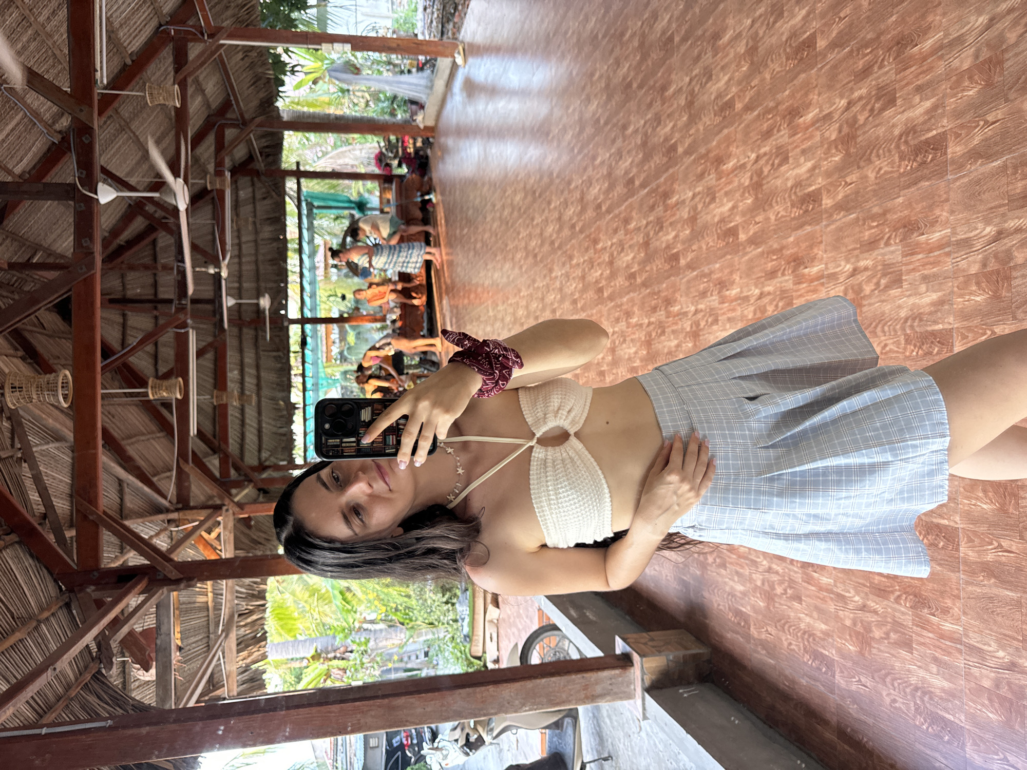 A young woman is taking a mirror selfie inside a wooden structure, potentially a yoga studio, in Mũi Né, Vietnam. The structure has a thatched roof supported by a wooden frame. Several ceiling fans are visible. Outside the structure, people are sitting on the floor practicing yoga. In the foreground, part of a scooter and other vehicles are visible, suggesting an urban setting. The woman is wearing a white knit top and a plaid skirt. She is holding a phone with a dark case. She is wearing a red beaded bracelet and a necklace. The floor is made of reddish-brown patterned tiles.