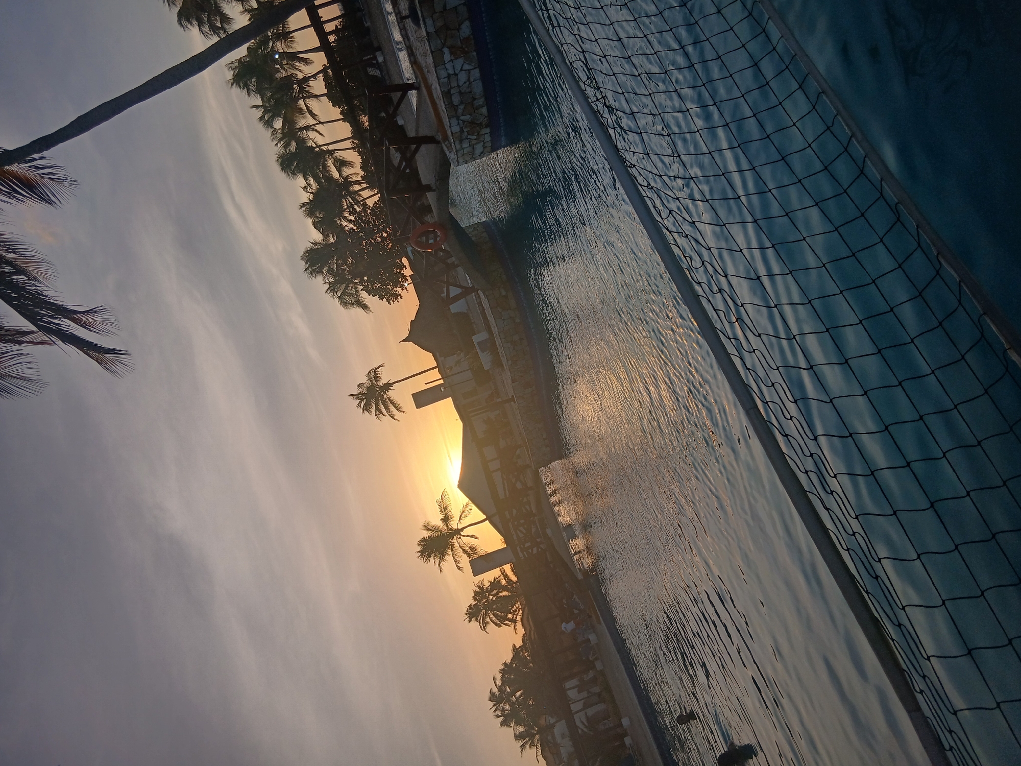 This image depicts a resort-style swimming pool complex in Altagracia, Venezuela, captured during either sunset or sunrise, with the light source low on the horizon and creating a backlit scene. The photograph is tilted diagonally.
The foreground features a black-netted structure, consistent with a water volleyball net, spanning across the pool. The expansive swimming pool water appears blue-green, with visible ripples and reflections of the warm, orange-yellow sunlight. Two individuals are partially submerged in the water on the left side, their heads and shoulders visible above the surface.
In the midground, a small, arched wooden bridge with railings crosses a section of the pool, supported by columns clad in light-colored stone. A red lifebuoy is affixed to the bridge railing. Beyond the pool, several buildings are visible, including one with a prominent pointed or thatched roof and others with shaded outdoor seating areas.
The background is dominated by numerous tall palm trees and other dense foliage, silhouetted against the sky, which transitions from bright orange and yellow at the horizon to a lighter grey-blue with faint cloud streaks overhead. Several indistinct figures are present in the shaded zones near the background structures.