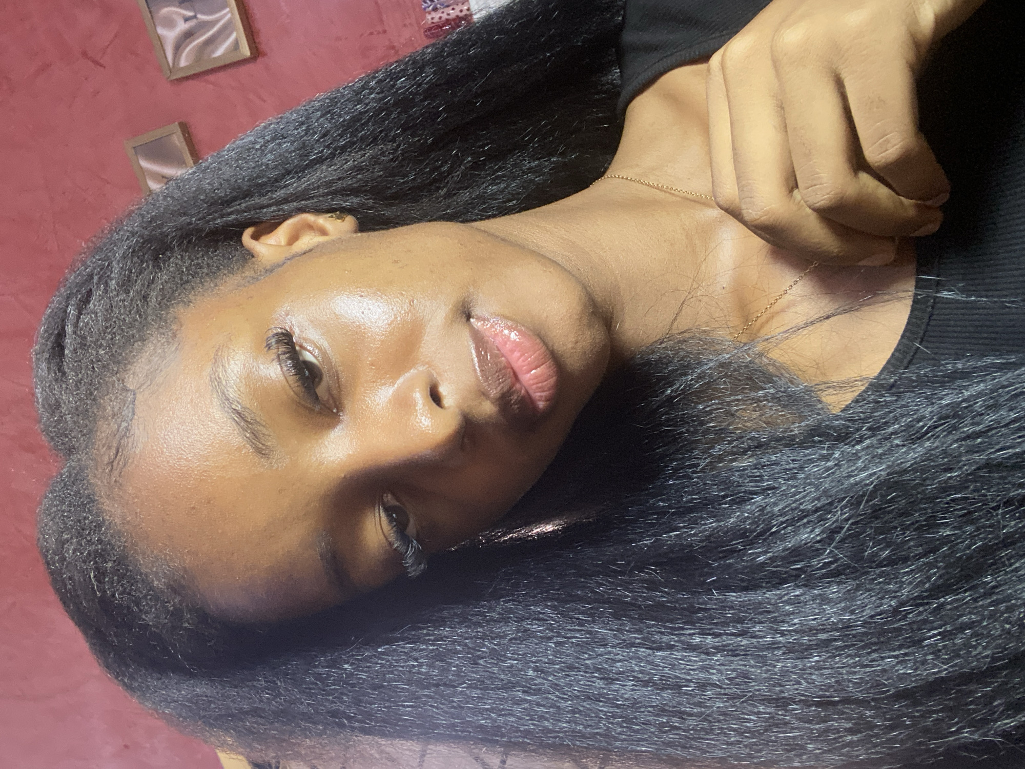 A young Black woman with long, dark, braided hair is shown from the chest up, angled slightly towards the camera. She is wearing a black top and a delicate gold chain necklace. Her skin has a dewy sheen, and she is wearing lip gloss. Her right hand is placed on her chest, near the necklace. The background is a textured reddish-pink wall with two small framed pictures. The scene appears to be indoors, possibly a bedroom or living space in Jos, Nigeria.
