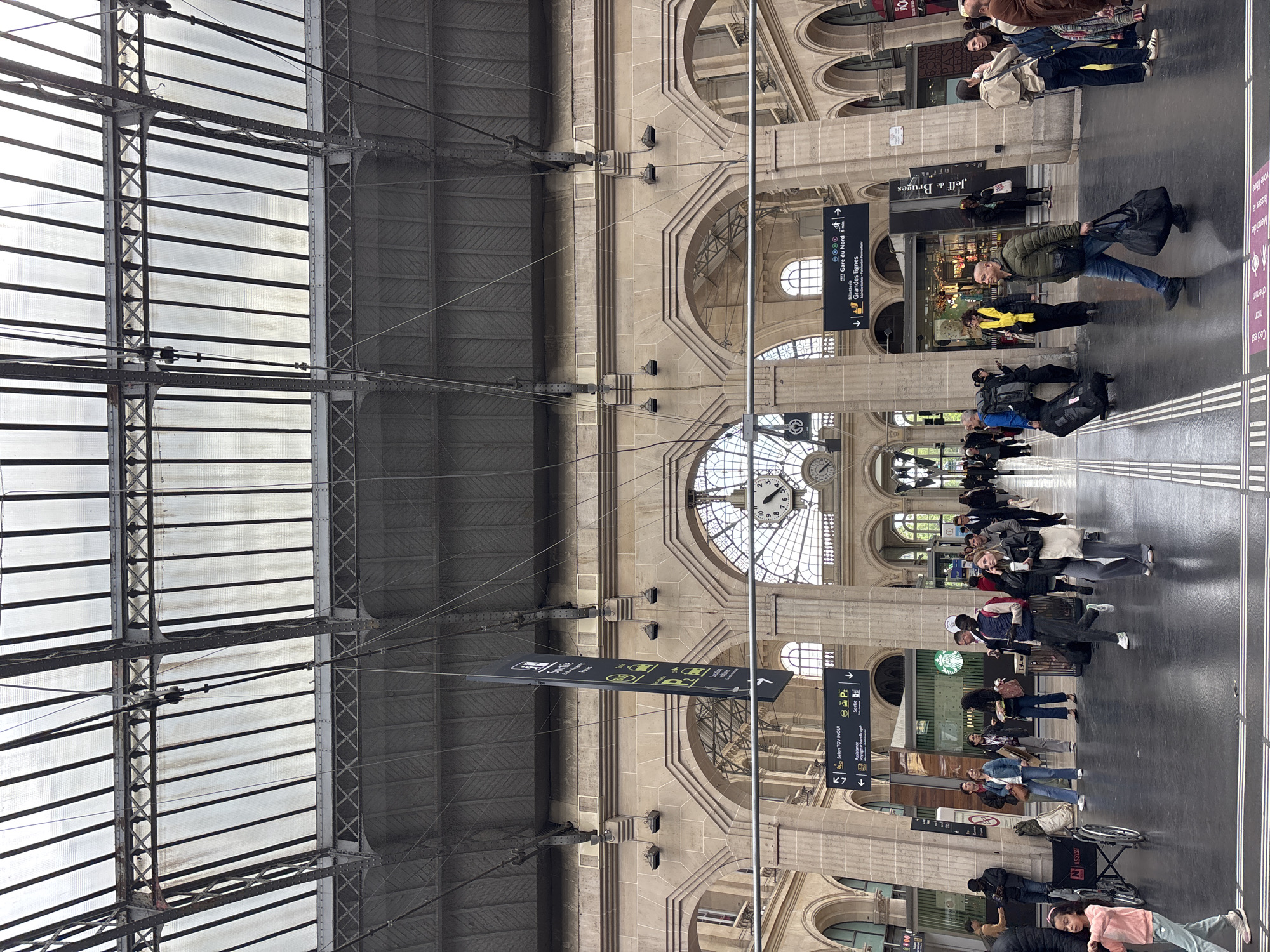 The image shows the Gare du Nord train station in Paris, France. The interior features a high, vaulted ceiling constructed of steel and glass, allowing natural light to illuminate the space. Several large clocks are integrated into the architectural design. Passengers are seen walking and waiting, some with luggage. Directional signs in French point towards various destinations and services. There are some commercial establishments. The people are wearing regular clothes.