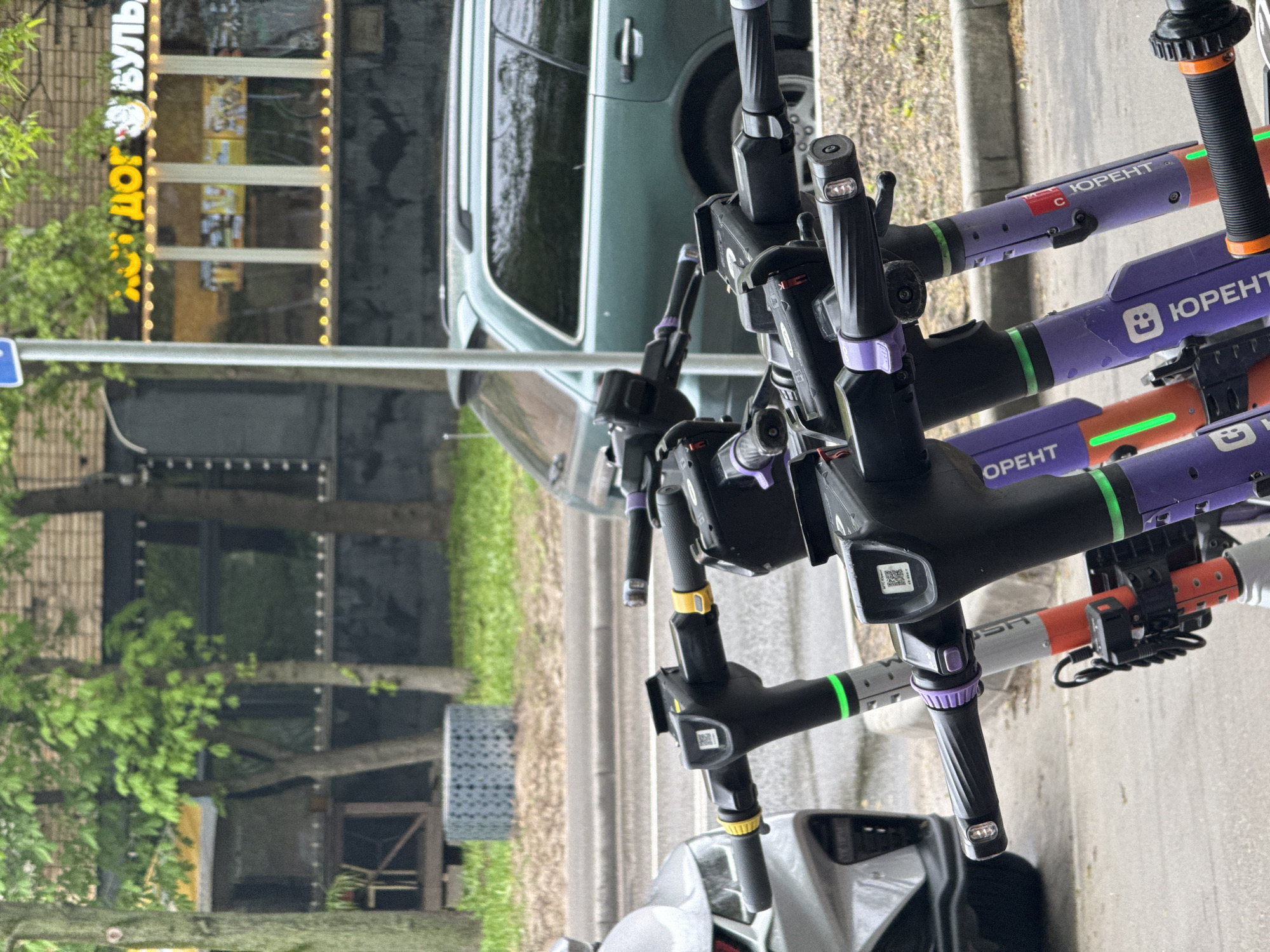 The image shows a group of parked electric scooters in Moscow, Russia. The scooters are branded "Юрент" (Urent), a local scooter rental company. The background includes a street, trees, a building with signs in Russian, and a parked car. The scooters are parked on the side of the road.