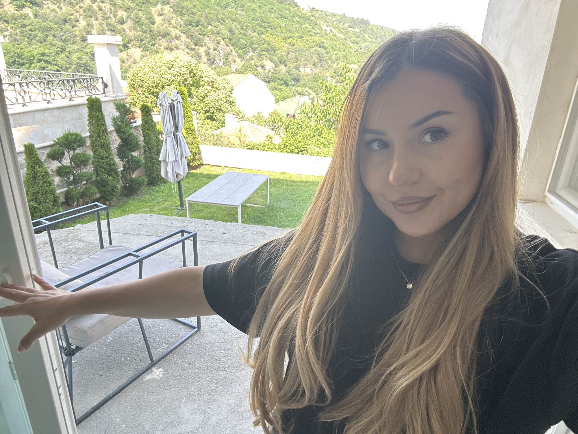 A female individual is depicted in the foreground, captured in a selfie-style photograph, positioned outdoors. She has long, light brown hair with blonde highlights, styled with a slight wave, and fair skin. Her eyes are light brown, and her lips are colored in a plum tone. She wears a black short-sleeved top and a delicate gold necklace with a circular pendant. Her right arm extends towards the left, with her hand touching a white vertical frame structure.
The background reveals a paved patio area. To the left, two modern outdoor chairs with dark metal frames and light grey cushions are visible. To the right of the chairs, a rectangular outdoor table with a light-colored top and dark frame is present. Beyond the patio, a green lawn extends, featuring several ornamental evergreen trees, including tall slender varieties and topiaries, as well as a large, folded, light grey patio umbrella. In the distance, multiple residential structures with tiled roofs are visible amidst abundant green trees and foliage. The far background is dominated by a steep, heavily wooded hillside or mountain range, characteristic of the natural landscape near Asenovgrad, Bulgaria. On the left side of the background, a portion of an elevated terrace with a stone balustrade and ornate dark metal railing is visible. The scene is brightly illuminated by sunlight.