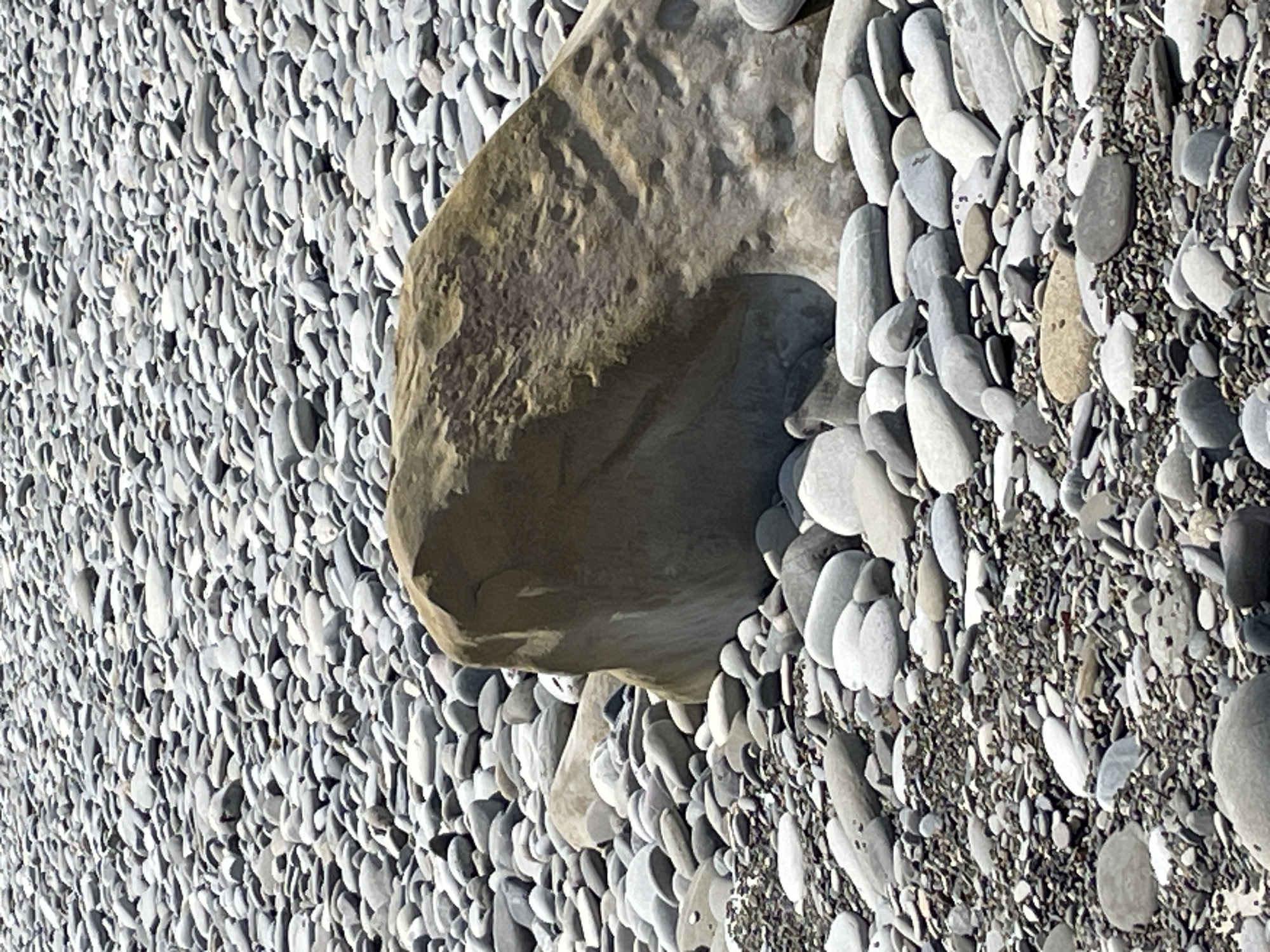 The image depicts a close-up, eye-level, horizontal view of a rocky beach. The foreground and midground are densely covered with numerous rounded pebbles and stones of varying sizes and shades of gray, white, and brown. A large, irregularly shaped, tan-colored rock with a rough texture is prominently positioned in the center of the frame, taking up a significant portion of the midground. The background consists of an even denser expanse of smaller, similar-colored pebbles, creating a textured, repeating pattern that extends to the horizon. The lighting suggests daylight, casting subtle shadows on some of the stones. There are no people or animals visible in the scene. The overall impression is one of a natural, unpopulated coastal environment.