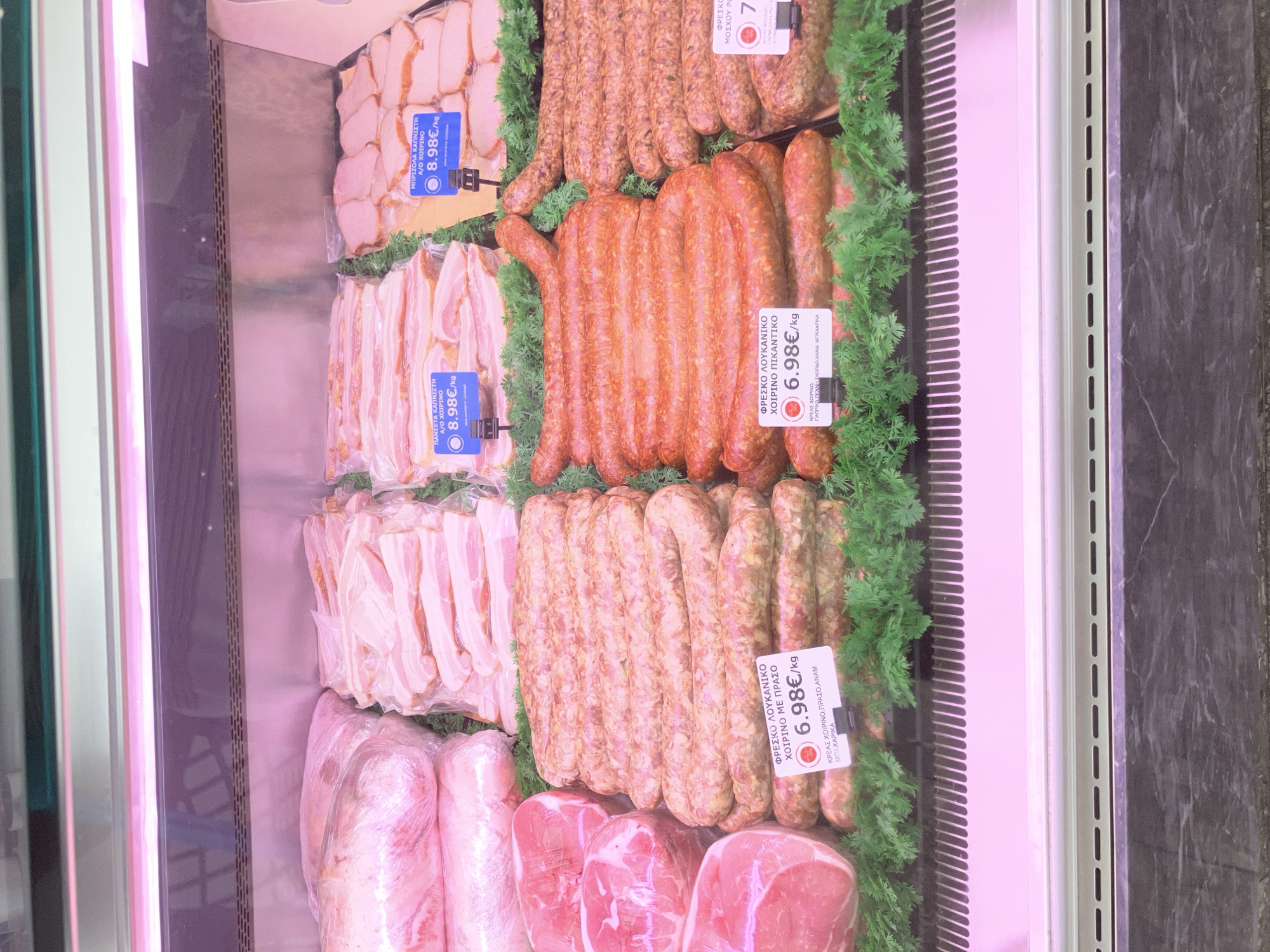 The image shows a refrigerated display case in a butcher shop located in Paleo Faliro, Greece. The case is filled with various cuts of meat, including what appears to be pork belly, sausages, and possibly pork loin. Several price tags are visible, indicating prices in Euros per kilogram for different meat products. The lighting is pinkish, characteristic of meat display refrigeration. There are no people visible in the image. The overall environment is clean and organized, suggesting a commercial establishment.