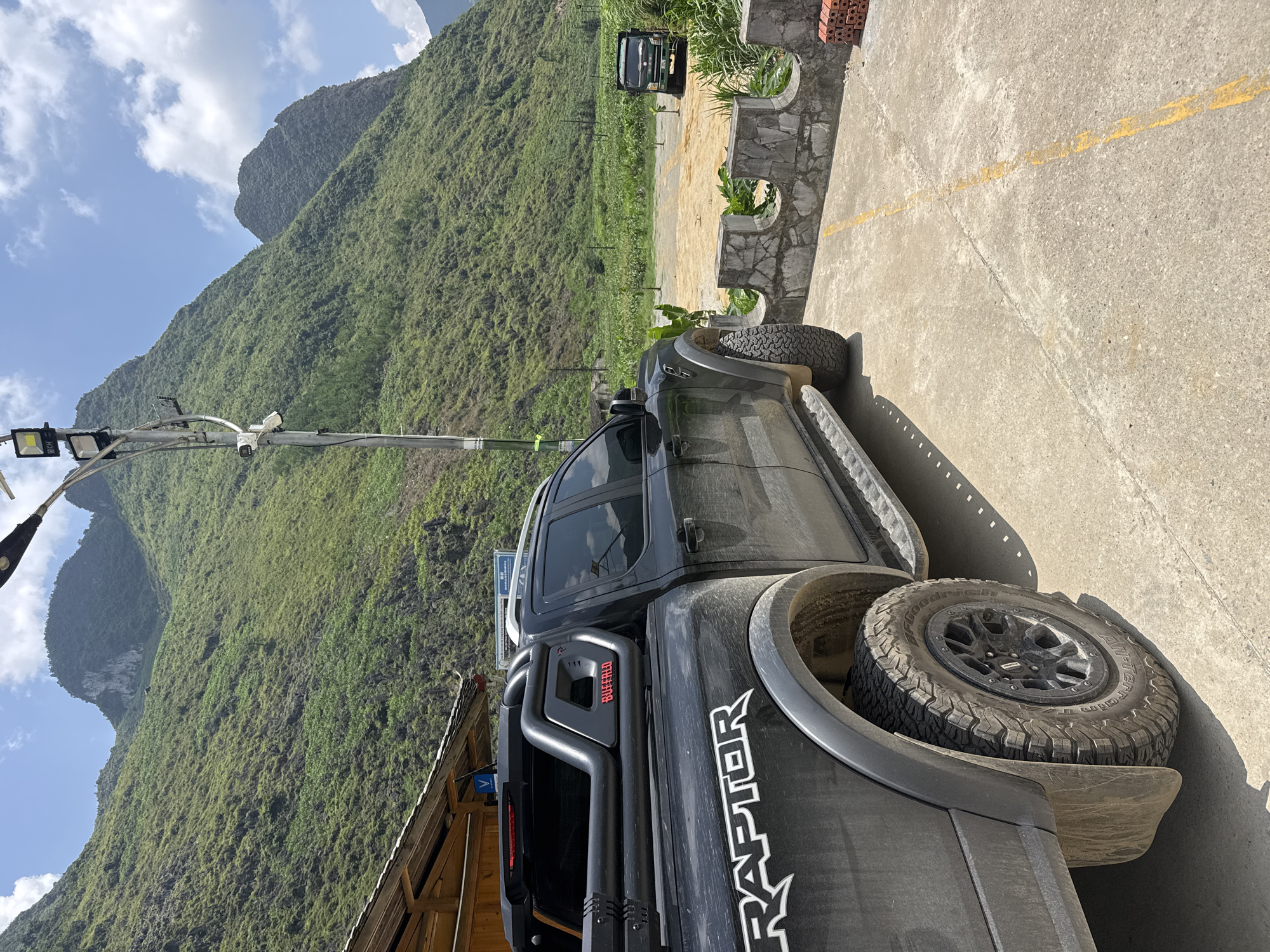 The image depicts a black Ford Raptor pickup truck parked near a rural area in Mèo Vạc Commune, Vietnam. The truck is dusty and has a spare tire mounted on the rear. The background features a large, green, mountainous landscape under a partially cloudy sky. A light pole with security cameras is visible to the left of the truck. A concrete wall with planted greenery and another vehicle parked further away can be seen in the background along the pavement.