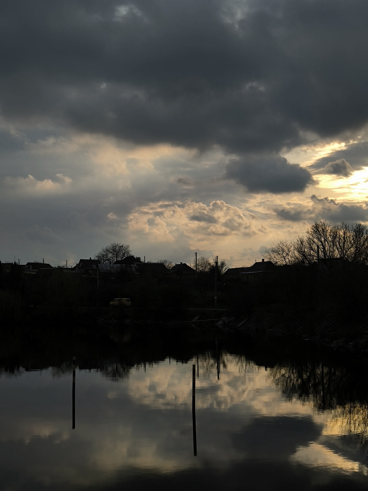The image shows a somber landscape, likely near a body of water, with a silhouette of a small village against a dramatic, cloudy sky.
**Environment:** The sky is dominated by dark, heavy clouds, with a patch of lighter clouds reflecting the setting or rising sun, giving the scene a melancholic atmosphere. Below, a body of water reflects the sky and silhouettes of trees and objects.
**Objects:** Silhouetted roofs of buildings and bare trees line the horizon, indicating a village or town. The foreground shows two utility poles.
**Location:** While the image does not explicitly confirm the location, the presence of Ukrainian style architecture suggests the image could be from a similar area, Vinnytsia, Ukraine.