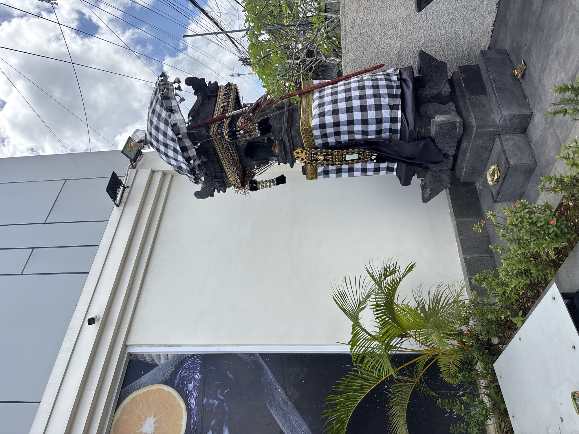 A partially obscured outdoor shrine, wrapped in a checkered cloth, is positioned in front of a modern building with an advertisement featuring produce. The shrine stands on a layered platform of dark stones. Tropical plants surround the shrine, including a small palm tree. Above, power lines stretch against a cloudy sky. A security camera is visible on the building's wall. The location is in Kuta Selatan, Indonesia.
