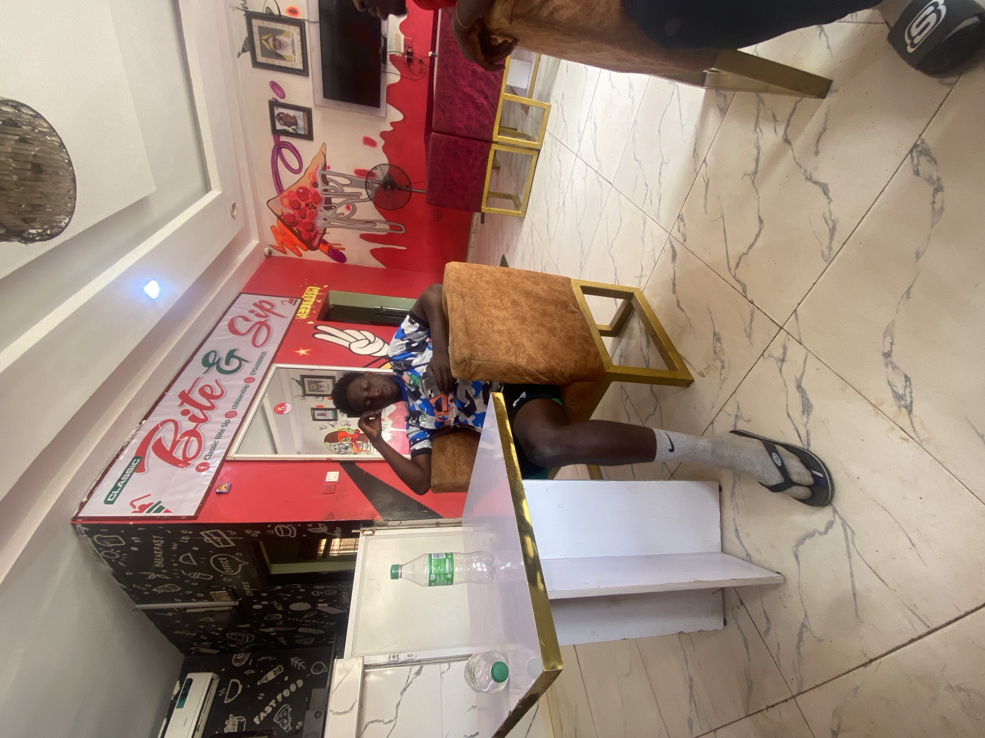 The image shows the interior of Classic Bite & Sip, a fast-food restaurant. A person is seated on a chair with brown cushions and a gold frame. The person is wearing a patterned shirt, shorts, and socks with sandals. A multi-tiered white shelf with a gold trim sits next to the person. A bottle of water is lying on the shelf. The restaurant has red and white walls decorated with food-themed artwork and a black and white section with food-related doodles. A television and framed photographs are mounted on the wall. There are burgundy-colored booth seats with gold-framed tables in the background. The floor is tiled with a marble pattern. The location appears to be Tukur Tukur, Nigeria, based on the OCR text "07040559025," which is a Nigerian phone number format.
