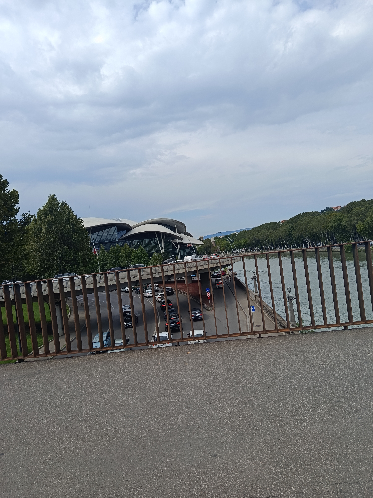 An eye-level, wide shot captures a bridge railing in the foreground, obscuring a view of a city street with moderate car traffic below, bordered by a river on the right and lush green trees on the left. In the background, a modern, architecturally distinct building with a white, curved roof sits amidst the trees. The sky is overcast with a mix of grey and white clouds.