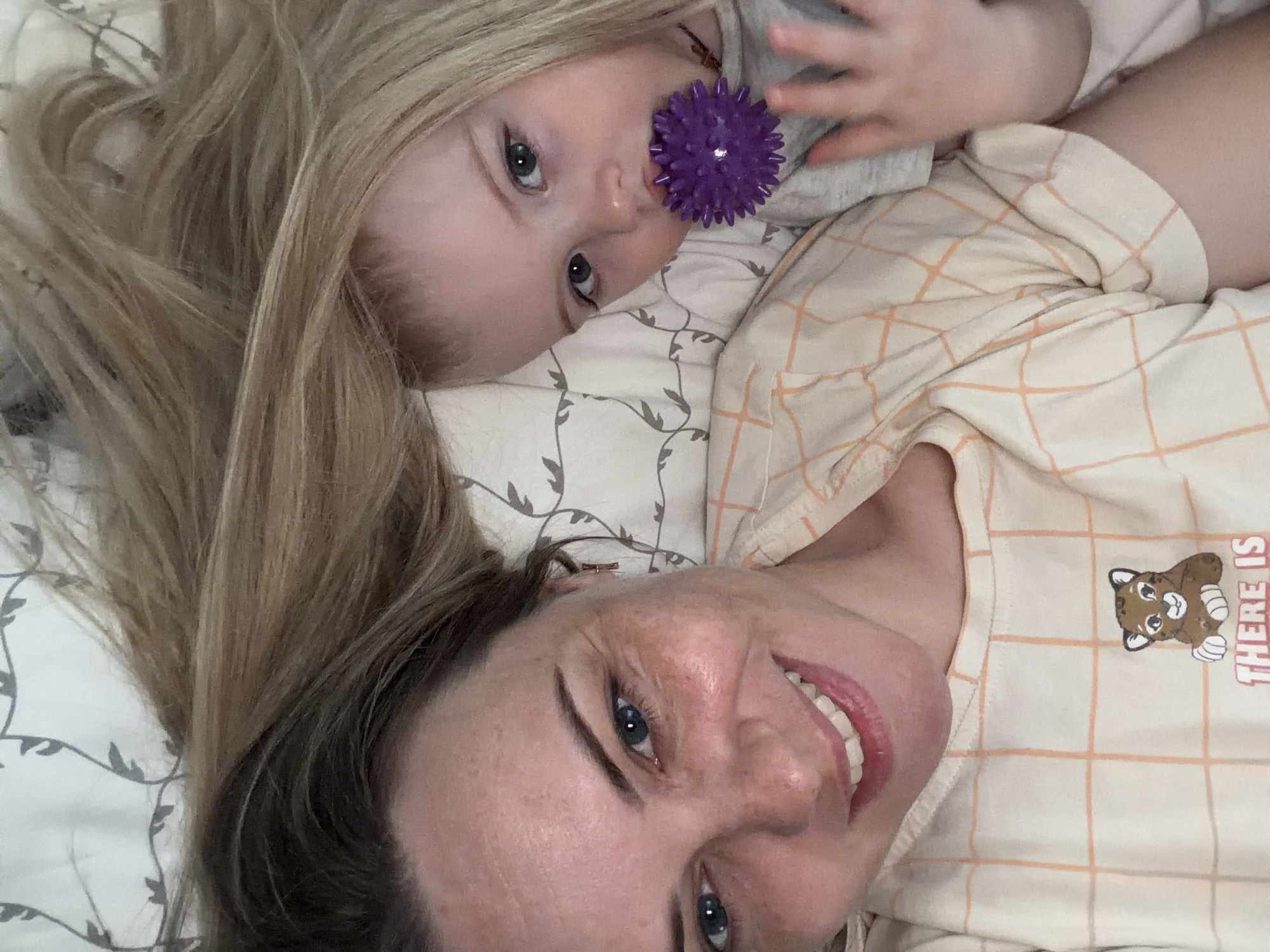 A selfie-style image depicts a woman with light brown and blonde hair smiling at the camera while lying on a bed with a baby. The baby, with blonde hair, lies perpendicular to the woman, partially overlapping her. The baby holds a purple textured ball near their face. The woman wears a light-colored plaid shirt with a cartoon bear graphic. The bed has a patterned sheet with a muted grey and white design. No city or location information is directly visible in the image to identify Votkinsk, Russia.