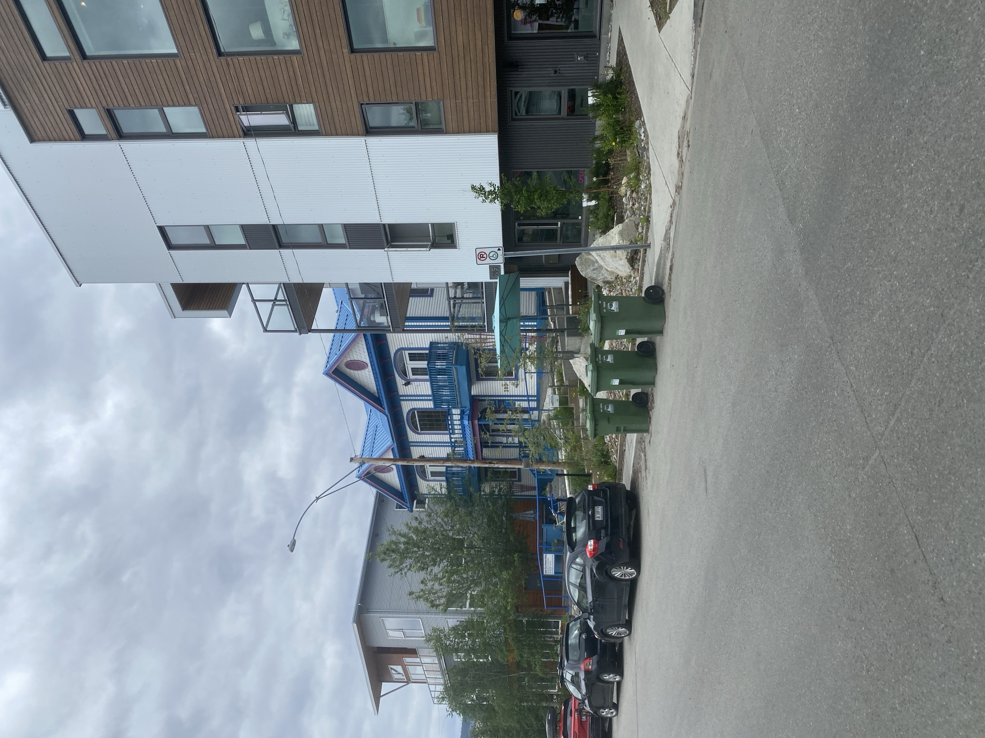 In Whitehorse, Canada, a street scene unfolds with a modern building dominating the right side, its facade a mix of brown wood paneling and white corrugated metal. A contemporary apartment building with balconies and large windows is visible on the left, behind a row of parked cars. Between these two structures, a colorful, older building with a bright blue roof and white trim stands out. The street is paved, with a sidewalk bordering the buildings. Several green garbage bins are placed along the sidewalk. A streetlight arches over the scene. The sky is overcast, suggesting a cloudy day. The overall impression is of a mixed-architecture urban environment.