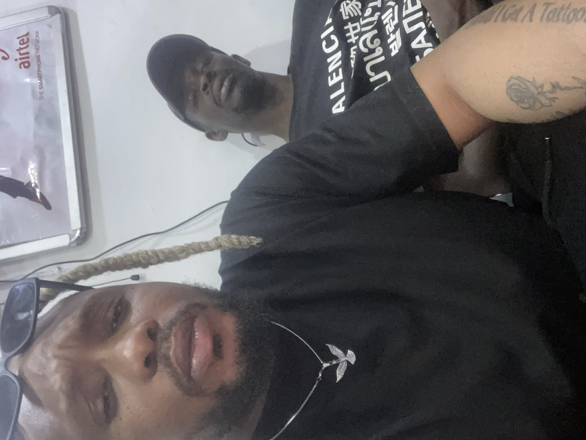 Two African men pose for a selfie indoors in Jos, Nigeria. The man in the foreground has braided blonde-tipped hair, sunglasses perched atop his head, a black shirt, and a silver bird pendant necklace. The man in the background wears a black baseball cap and a black t-shirt with non-English text. An "Airtel" advertisement is visible in a frame behind the men. The right arm of an individual with a tattoo is partially visible in the upper right.