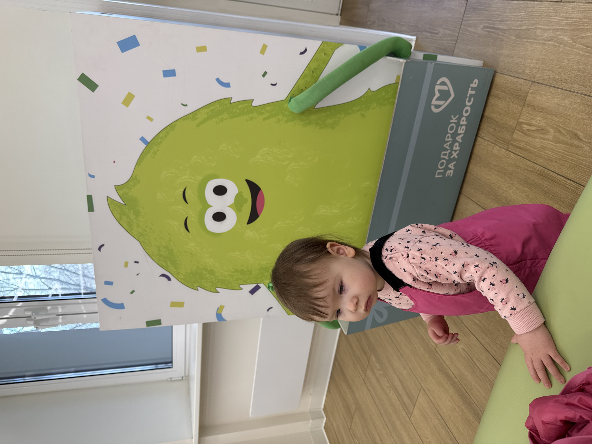 A young child is positioned near a large cardboard cutout of a green monster with a smiling face, located inside a room. The cutout sits atop a gray rectangular box with Russian text "ПОДАРОК ЗА ХРАБРОСТЬ," which translates to "GIFT FOR COURAGE." The child is wearing a pink outfit with a floral pattern top and a pink overall dress. The room has light-colored walls, a wooden floor, and a window. The location could be Moscow, Russia, given the Russian text.