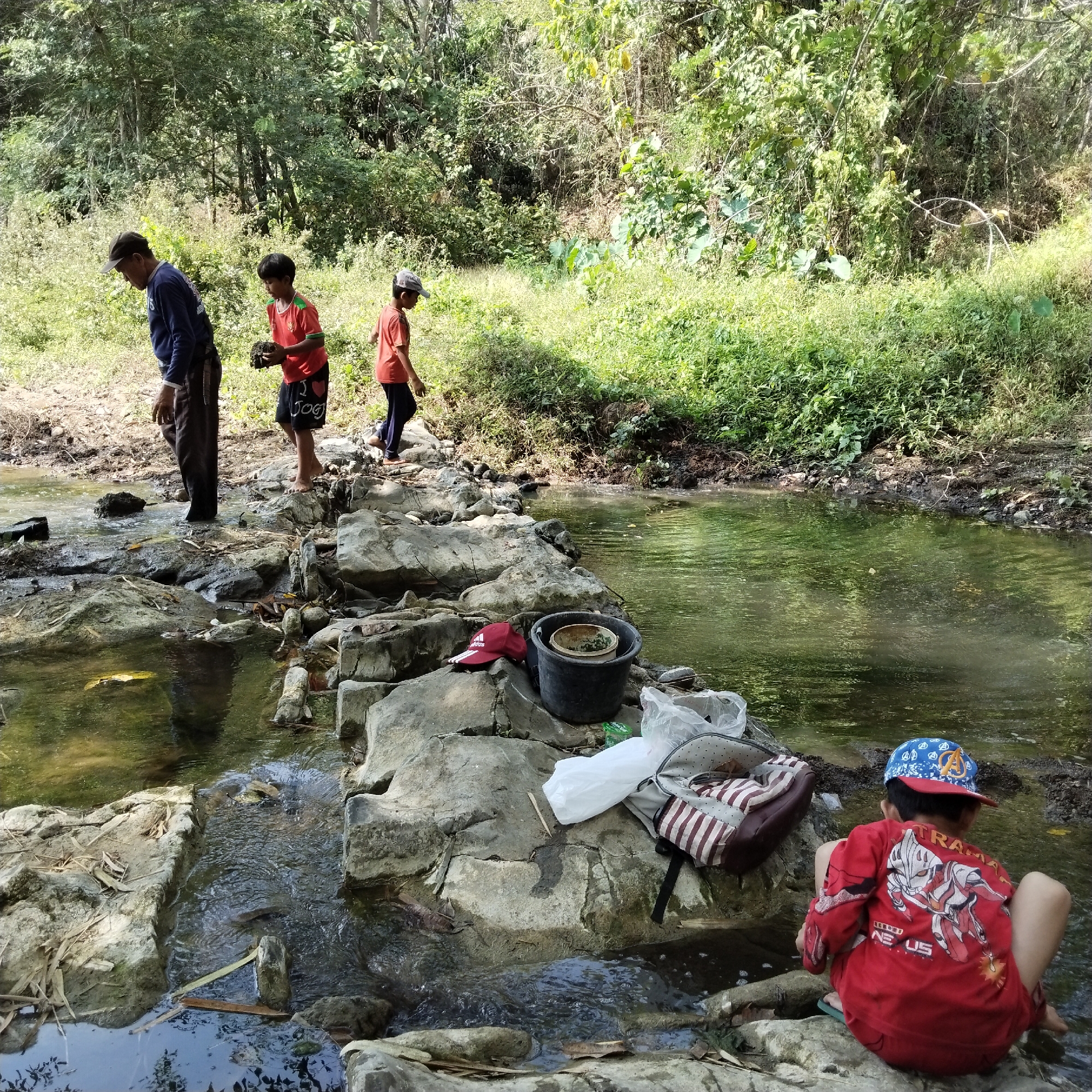 In Karangbaru, Indonesia, a group of four individuals are seen by a shallow, rocky stream in a wooded area. The stream flows over a bed of stones and pebbles. On the left bank of the stream, an adult male wearing a blue long-sleeved shirt and dark pants stands near the water's edge, facing away from the camera. To his right, a child in a red shirt and black shorts holds a dark, irregular object. Further to the right, another child in an orange t-shirt and dark pants walks away from the viewer. On the right bank, a child wearing a red long-sleeved shirt with graphics and a blue baseball cap is crouched on rocks, facing the stream. Several items are scattered on the rocks near the stream, including a red cap with a white logo, a dark bucket with green contents, a white plastic bag, and a backpack with red and white stripes. The surrounding environment is dense with green vegetation, including trees and bushes, indicating a natural, undeveloped area. The sunlight suggests it is daytime.