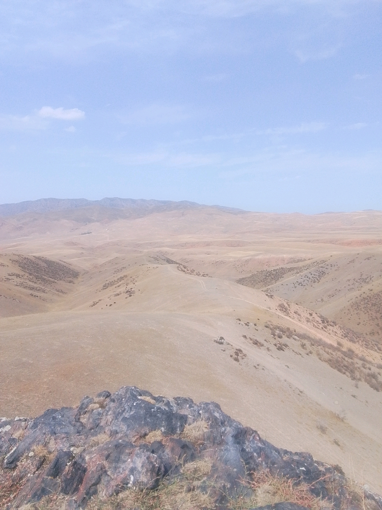 A wide, high-angle shot captures a vast, arid landscape under a pale blue sky with scattered, wispy clouds. The foreground features a cluster of dark, jagged rocks, interspersed with dry grasses. Beyond the rocks, the terrain unfolds in rolling hills and valleys, covered in dry, tan-colored vegetation. The midground shows more detailed contours of these hills, with some areas appearing more densely vegetated with scrubby bushes. In the distance, a range of muted, hazy mountains stretches across the horizon, suggesting a considerable expanse. The overall impression is one of a remote, undeveloped, and dry environment, characteristic of a steppe or mountainous desert region. The scene is devoid of human presence, animals, or any structures, emphasizing the natural and desolate character of the location. The lighting suggests it is daytime, possibly morning or late afternoon, given the soft, diffused light. The specific location in Maydan, Kyrgyzstan, indicates a region known for its natural landscapes, potentially high altitude and semi-arid climate.