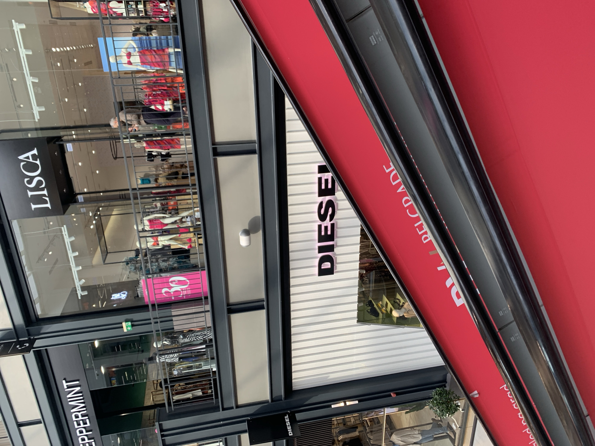 This image captures a section of a modern shopping environment, likely an indoor mall or a covered commercial street, featuring several retail stores. The perspective is angled, looking across and down into the shopfronts.
In the foreground, a prominent red awning or signage structure dominates the right side of the frame, with reflective metallic elements. Visible text on this red surface, written vertically, spells out "BELGRADE".
To the left and further back, several store signs are clearly visible. The store "LISCA" is prominently displayed on a black background. Below it, the store "PEPPERMINT" is also visible on a black sign. Another store, "DIESEL", is identifiable by its name written in black letters on a white, ridged facade, and also on a smaller black sign below it.
Behind the glass windows of the shops, various mannequins display clothing, including swimwear in red and pink hues. Inside one shop, a "30%" discount sign is noticeable in pink. The interior spaces feature bright overhead lighting. One person, an older man in a light jacket, can be seen inside a store, blurred in the background. The architecture features dark metal framing and large glass panels, giving a sleek, contemporary feel. A small potted plant adds a touch of greenery near the bottom of the frame. The overall scene suggests a vibrant retail atmosphere in Belgrade, Serbia.