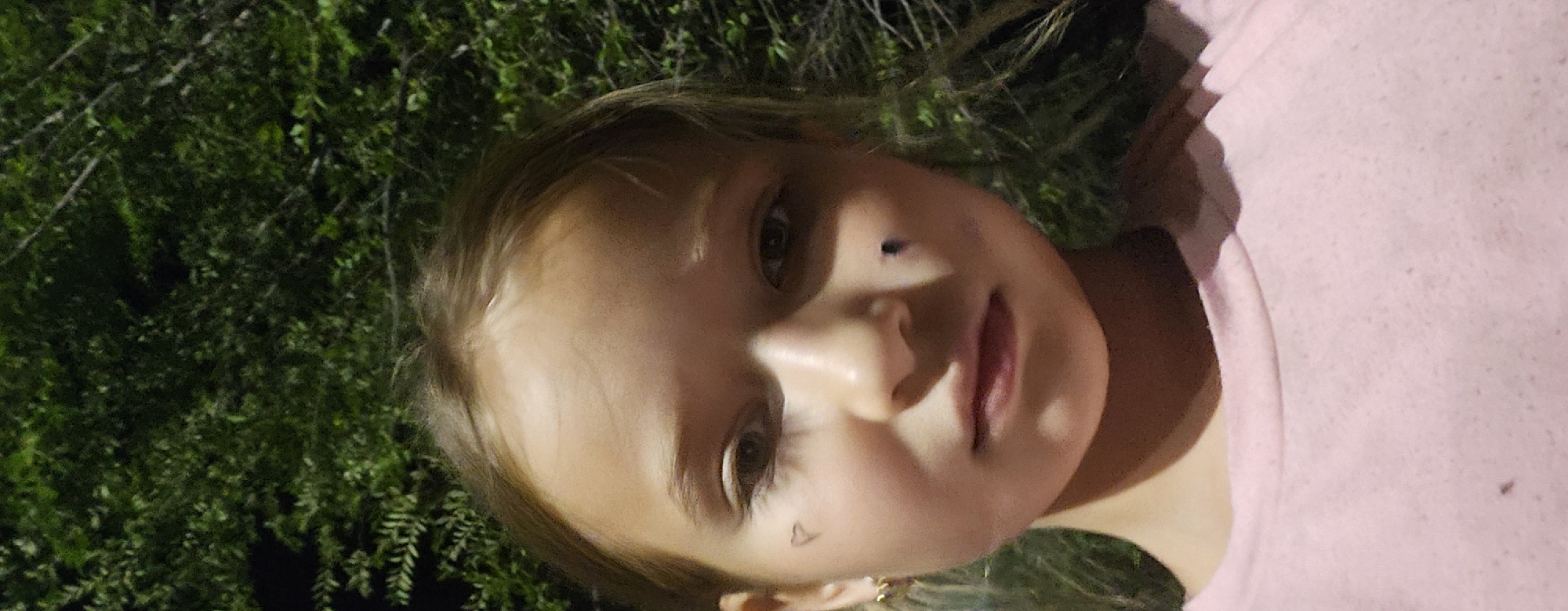 This is a close-up, portrait-style photograph of a young girl. She has blonde hair, fair skin, and is looking directly at the camera. Her eyes are wide, and her mouth is slightly open, giving her a curious or surprised expression. There is a small black dot on her cheekbone and a faint outline of a heart shape on her jawline, which could be temporary markings or drawn on. She is wearing a pink t-shirt. The background is filled with dark green foliage, suggesting she is outdoors, possibly in a garden or park. The lighting is somewhat dramatic, with strong highlights and shadows on her face, indicating it might be evening or a dimly lit area. There is no visible text in the image. The provided location context is Semey, Kazakhstan, but there are no visual cues within the image itself to confirm this location.