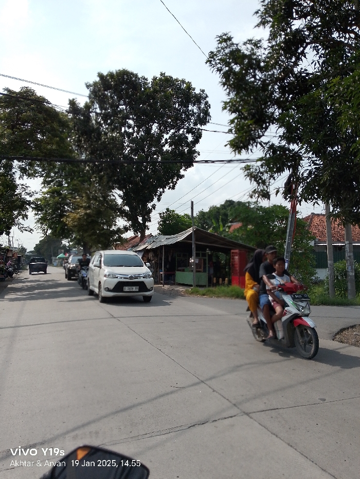 This full-shot image captures a sunny daytime street scene in Arjawinangun, Indonesia. The asphalt road, showing some cracks and lines, occupies the foreground and center.
In the mid-ground, a white multi-purpose vehicle (MPV), likely a Toyota Avanza, is prominently featured, facing towards the camera and slightly to the left. Its license plate, "B 5656 BV," is visible. Further down the road behind the white car, other vehicles, including a dark-colored car and several motorcycles, are visible, suggesting moderate traffic.
On the right side of the frame, a moped carries three people: an adult male driving, an adult female riding pillion, and a child seated between them. The moped is red and white, and its license plate reads "E 6431 BY." They appear to be in motion, possibly turning or driving along the road.
The background features several roadside stalls or small shops with simple structures, characterized by corrugated metal roofs and exposed frames, situated on the right. Beyond these stalls, the red-tiled roofs of houses and lush green trees are visible under a bright, partly cloudy sky. More dense foliage and trees line both sides of the road, and overhead power lines crisscross the sky.
The weather is bright and sunny, indicated by the strong lighting and visible shadows. A timestamp in the bottom-left corner reads "19 Jan 2025, 14.55," confirming the mid-afternoon time. Other visible text includes "vivo Y19s" and "Akhtar & Arvan," likely camera metadata. The scene depicts a common daily activity of transportation and commerce in a local Indonesian setting.