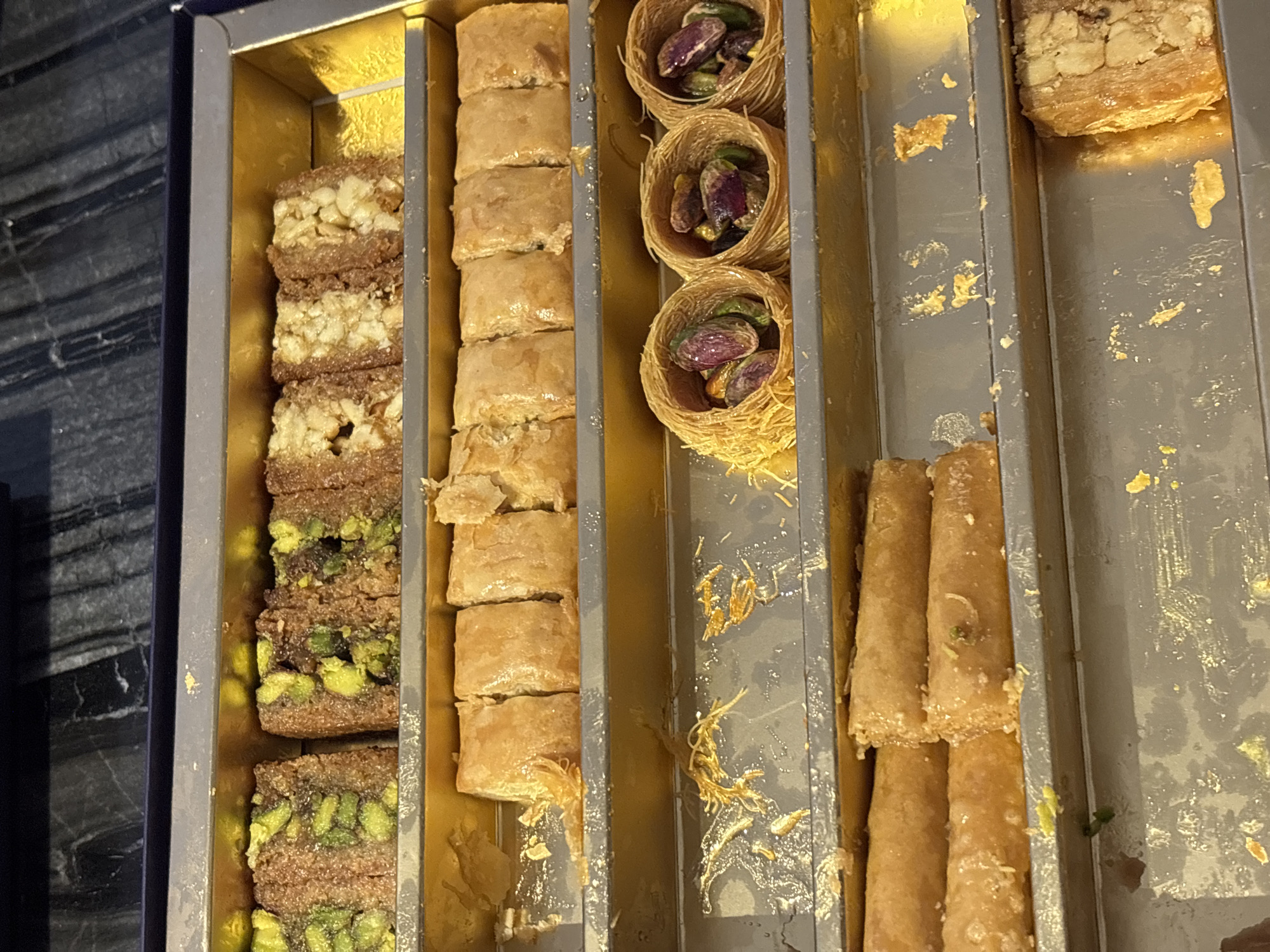 A top-down, medium shot captures a collection of Middle Eastern pastries arranged in a rectangular, multi-tiered golden box. The pastries exhibit a variety of shapes, textures, and fillings. The top tier features rectangular pieces of baklava-like sweets, some studded with pistachios and others with nuts. The middle tier presents rolls of phyllo dough, some filled with nuts and pistachios, and nests of shredded phyllo dough filled with pistachios. The bottom tier displays elongated, cylindrical pastries, likely also baklava, alongside a single rectangular pastry filled with nuts. The box is placed on a dark, possibly marble, surface. This image is not specific to Dubai, United Arab Emirates.