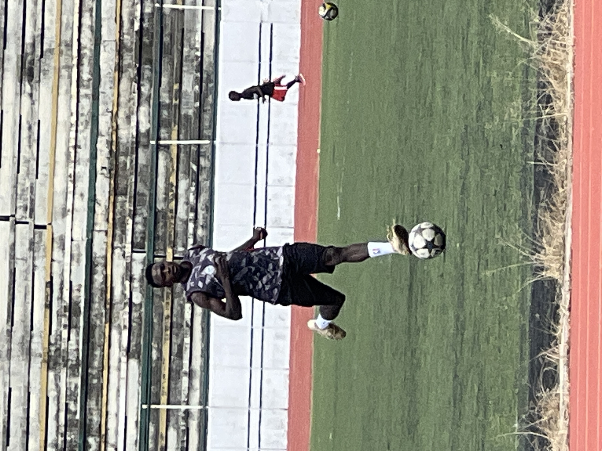 The image captures a dynamic scene on a football pitch, likely within Rafin Par, Nigeria, as suggested by the provided context. The foreground is dominated by a young man in mid-stride, dribbling a soccer ball. He is wearing a sleeveless, patterned jersey and black shorts, with white socks and athletic shoes. His focus is intently on the ball as he moves across the green turf. In the middle ground, another individual, also a male, is running in a similar direction, dressed in a black t-shirt and red shorts. The background reveals the tiered seating of a stadium, characterized by weathered concrete and faded yellow and green horizontal stripes. The overall environment suggests an outdoor training session or practice match.