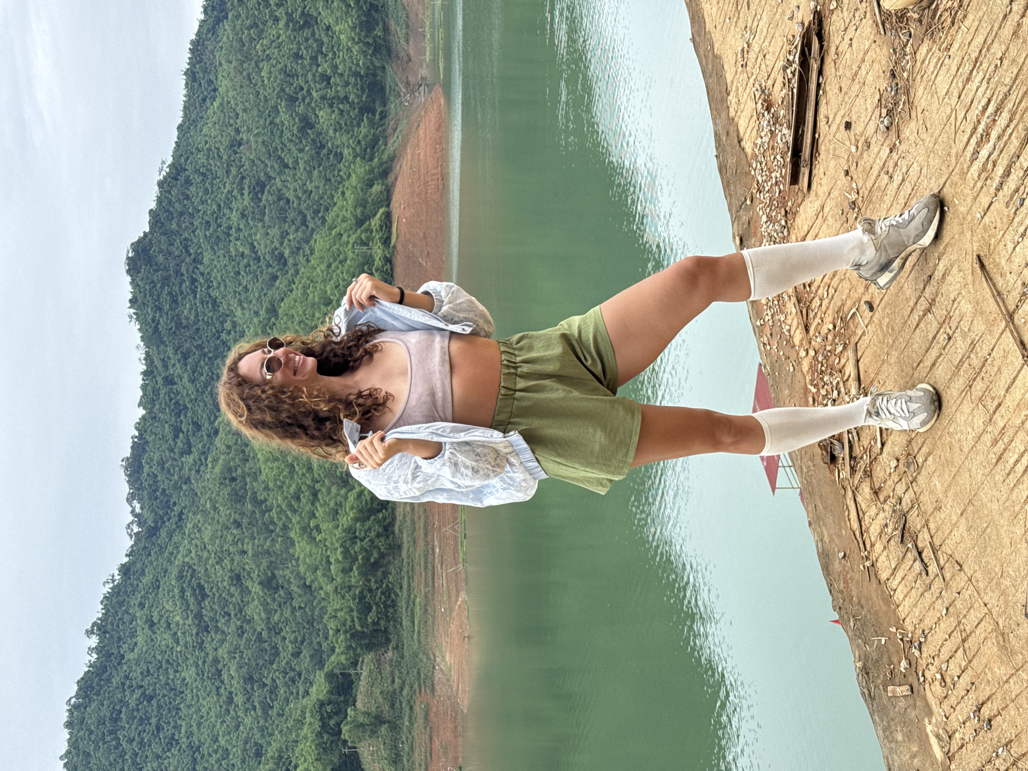 A young woman with curly brown hair, wearing sunglasses, a light blue jacket over a pink top, green shorts, white socks, and gray sneakers, poses on a concrete surface. Behind her, a vast, calm, green lake is surrounded by lush, tree-covered mountains under a cloudy sky. The scene appears to be located near Bản Phúc, Vietnam, a region known for its natural beauty and large bodies of water. The concrete area where the woman stands is rough and somewhat deteriorated, with scattered debris.