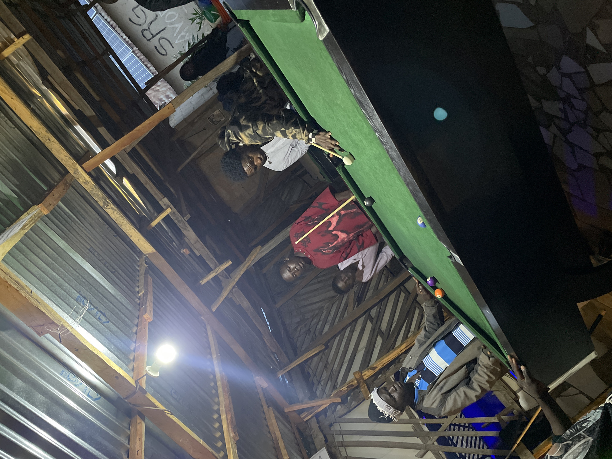 The image depicts an indoor scene with several individuals engaged in a game of billiards. The environment appears to be a casual establishment, possibly a bar or recreational center, with a corrugated metal roof supported by wooden beams. Natural light seems to be limited, with a bright overhead bulb illuminating parts of the scene.
In the foreground, on the right side of the frame, a person wearing a patterned hooded jacket is actively playing billiards, aiming with a cue stick at a white ball on a green felt table. Another individual, dressed in a white shirt, stands behind the player, observing the game. To the left, a person wearing a grey jacket over a blue and white striped shirt and a white headwrap is positioned near the billiard table, also seemingly participating or watching. Further back, another person in a red shirt is visible. The billiard table is set with several colored balls, including an orange ball and a purple ball.
The background reveals a rustic interior with wooden paneling and what appears to be some solar panels visible on the roof outside. Graffiti with text, including "SRG MAYO," is present on a wall. The overall impression is a lively, social gathering centered around the game. The specific location is identified as Durimi, Nigeria.