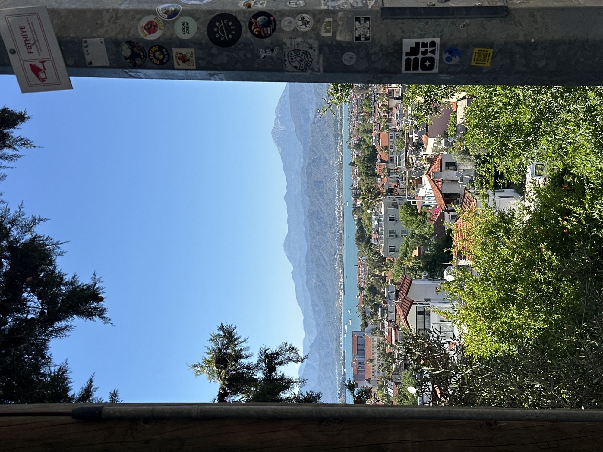 The image displays a panoramic view of a coastal town, a bay, and mountains, captured from an elevated perspective. It is currently rotated 90 degrees clockwise.
In the foreground, a dark wooden surface is visible at the bottom, topped by a metal railing. This railing is heavily adorned with numerous stickers. Prominently visible among them is a white rectangular sticker with a red graphic (resembling a stylized boat or hook), featuring the text "FETHIYE" in red capital letters, with smaller, less legible text below it. Other stickers include "LOCAL CREW", "EAT DOG GROW", "ABCDE", "ALOHA TEAM", "ALWAYS", and various abstract designs and symbols. Dark green coniferous trees are seen framing the view from the lower left and upper left corners (as if correctly oriented).
The midground features a vibrant blue body of water, likely a bay or the sea, stretching horizontally across the scene. Several small white boats are dotted across the water. Along the shoreline and extending up the gentle slopes, a densely packed town is visible, consisting of numerous buildings with predominantly white facades and terracotta-colored tile roofs. Lush green trees and bushes are interspersed throughout the urban landscape.
In the background, a series of large, hazy blue-gray mountains rise majestically, forming a dramatic backdrop to the coastal settlement and extending along the horizon.
The sky above is clear and bright blue, indicating sunny weather. The scene is bathed in bright daylight, suggesting it is midday or early afternoon. There are no people visible, and no specific activity is taking place; it is a static landscape view of Fethiye, Turkey, as indicated by the sticker.