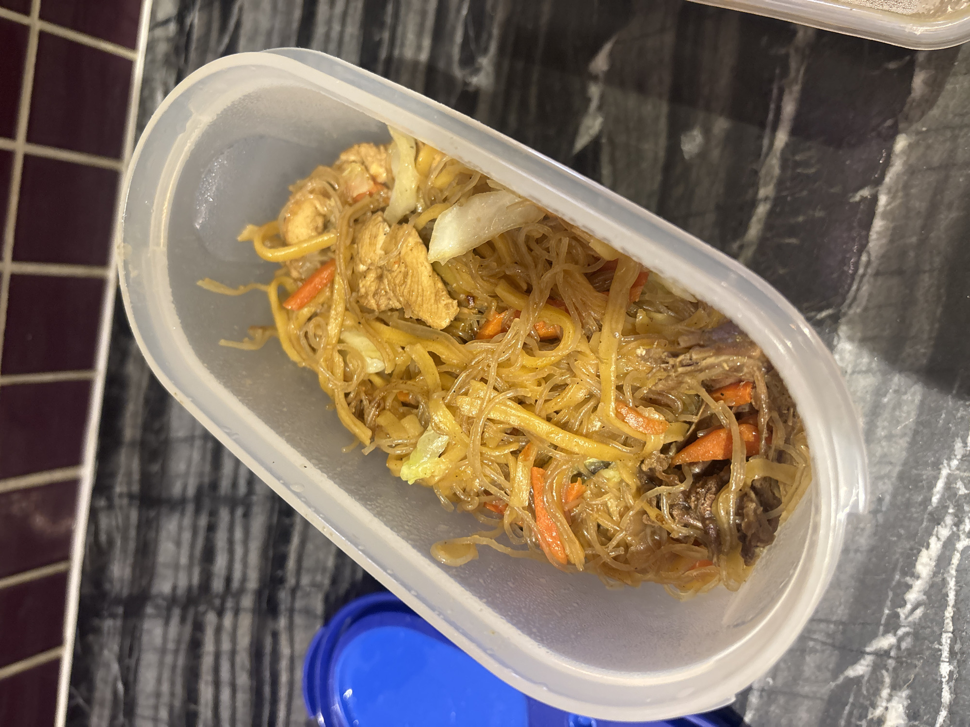 A transparent, oval-shaped plastic food container is filled with a noodle dish that contains shredded chicken, beef, and julienned carrots. The dish appears to be stir-fried with a light brown sauce. The container is placed on a dark gray countertop with a marbled pattern. To the left of the container, a blue plastic lid is partially visible. The background shows a backsplash with small, dark purple square tiles. The overall lighting suggests an indoor setting, possibly a kitchen. The scene does not provide any specific clues to pinpoint its location in Dubai, United Arab Emirates.