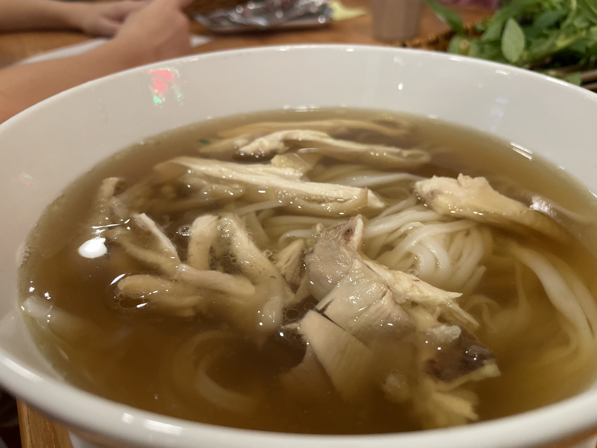 A close-up shot shows a bowl of pho. The bowl is white and filled with a light brown broth. Noodles and pieces of white meat, likely chicken, are submerged in the broth. Small bubbles and droplets of oil are visible on the surface of the soup. In the background, out of focus, are glimpses of what appears to be a dining setting. A wooden table is visible, and to the left, a person's arm and hand are slightly blurred, suggesting they are seated at the table. To the right, a small bunch of green herbs, possibly basil or mint, is also out of focus. The lighting suggests it might be daytime, indoors, in a casual eatery. The image captures a moment of someone enjoying a meal. No visible text is present in the image. The context provided suggests the location is Thủ Đức, Vietnam, a place known for its Vietnamese cuisine, including pho.