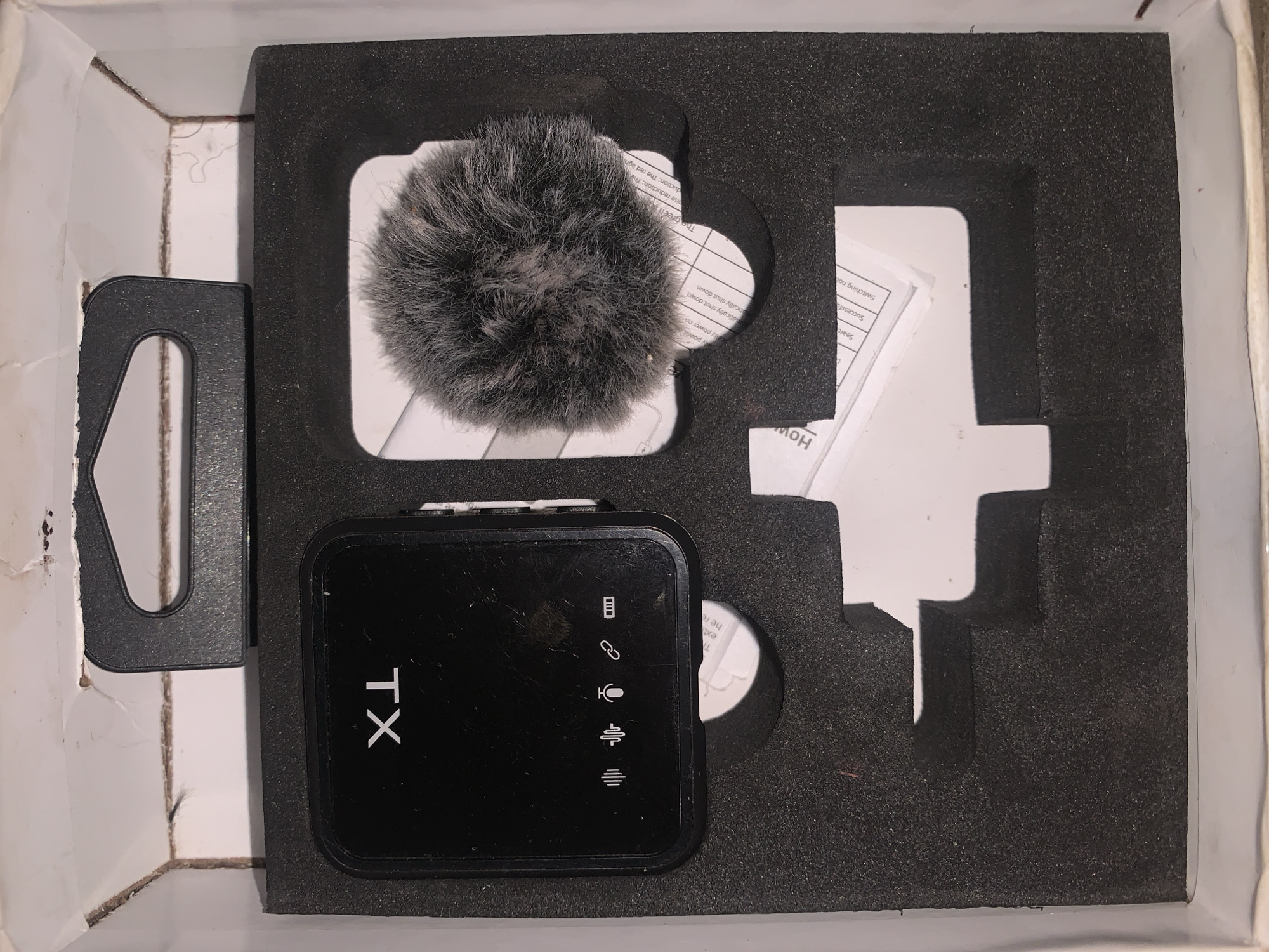 A black wireless microphone transmitter with "TX" printed on it is positioned in a black foam cutout within a white cardboard box. To the right of the transmitter is a furry windscreen. Below the transmitter is a small circular object with text printed on it. In the bottom right of the box is another white cutout with a paper document peeking out. The box is situated in an unknown location.