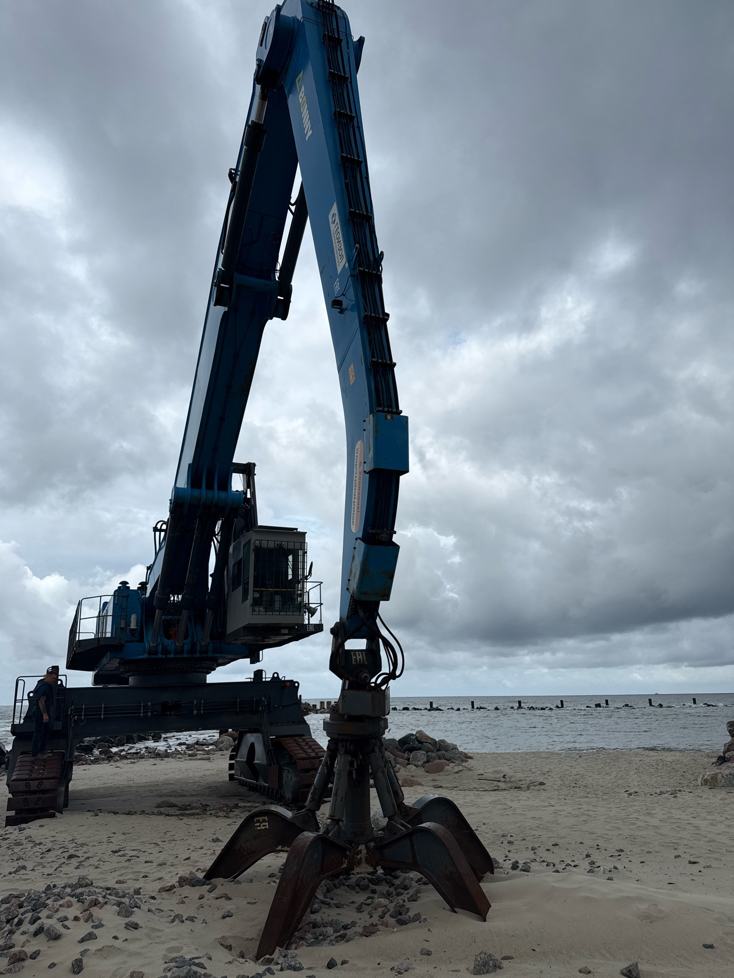 An industrial grabber crane is situated on a sandy beach with scattered rocks in Svetlogorsk, Russia. The crane is blue and features a large, articulated arm with a claw attachment at its base. The crane's cabin is visible, and it appears to be positioned on tracks. The background shows a body of water, likely the Baltic Sea, with a line of wooden posts extending into it, suggesting a breakwater or remnants of a pier. The sky is overcast with heavy, grey clouds. A single person, partially visible, is standing on the tracks of the crane. The overall scene depicts industrial equipment on a coastal environment under a cloudy sky.