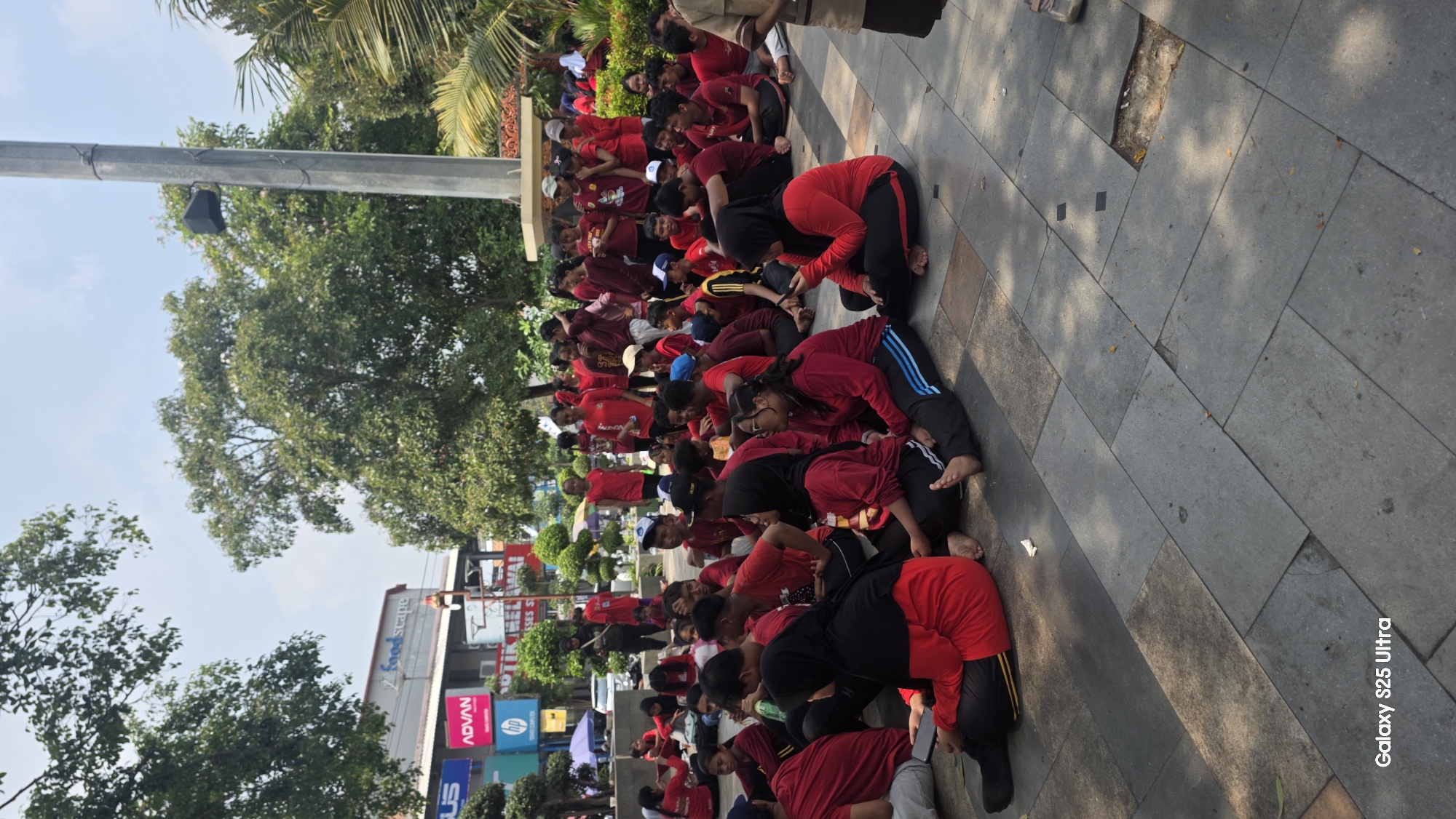 A large group of young people, likely students, are seated on a tiled outdoor plaza in front of a building displaying various commercial signs. The individuals are predominantly wearing red tops, with some wearing black bottoms and head coverings. They appear to be gathered for an event or activity, with many looking towards the camera or engaged in conversation with each other. The background includes trees, a tall pole with a speaker, and a clear sky. The overall scene suggests a public gathering in an urban setting.