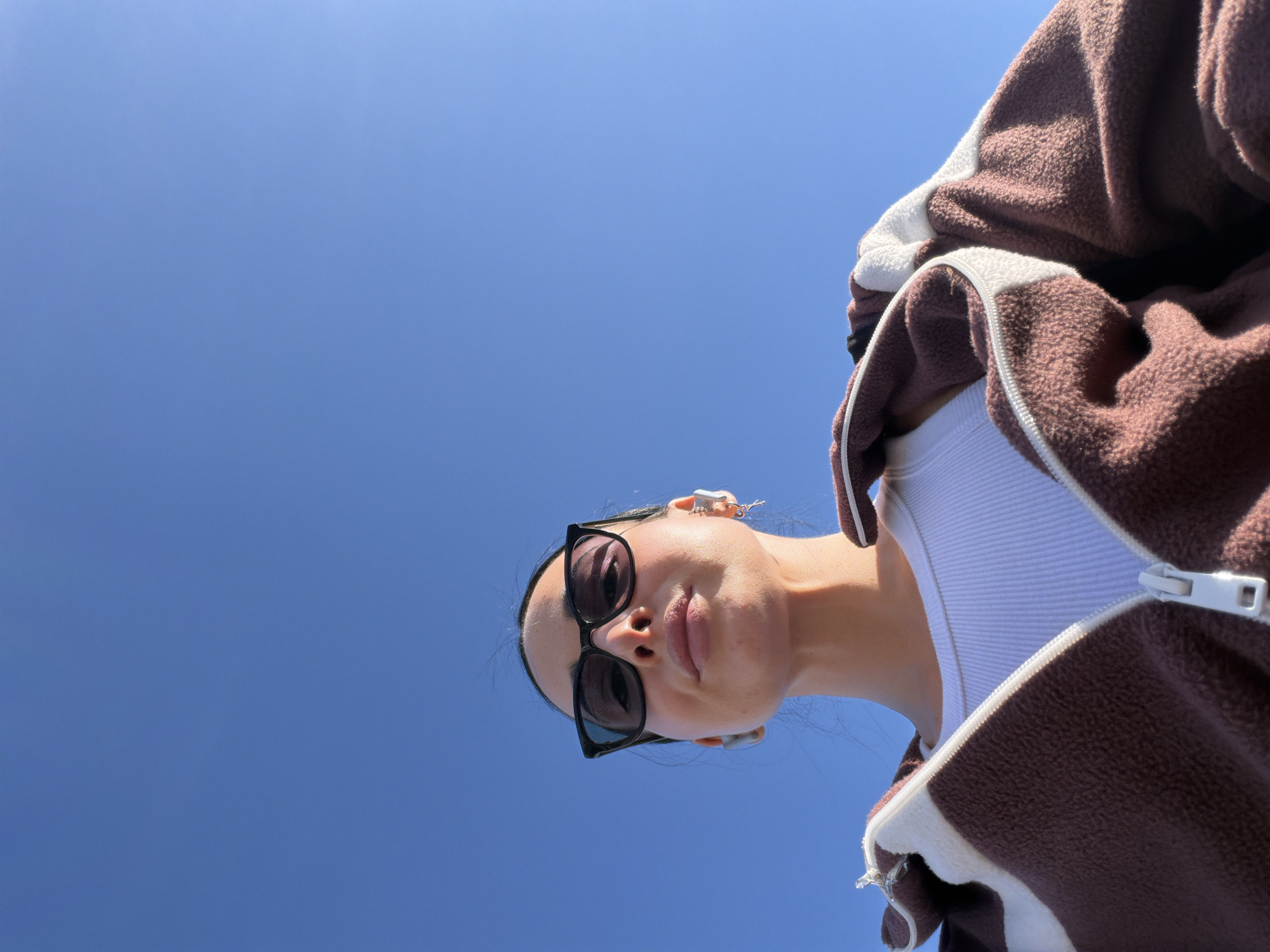 A low-angle close-up captures a person looking directly down towards the camera, framed against a clear, bright blue sky. The subject wears black, rectangular-framed sunglasses and has their lips slightly parted. Visible accessories include small, silver earrings, one of which appears to be shaped like an animal, possibly a frog, on the upper ear. The person is dressed in a brown and white fuzzy or fleece zippered jacket, with a white ribbed tank top visible underneath. The jacket features a white zipper and white accents. The strong, direct sunlight creates distinct highlights and shadows on the person's face and clothing, indicating a sunny day, likely during midday or early afternoon. The sky in the background is uniformly blue with no clouds. This scene is set in Saint Petersburg, Russia. There is no visible text in the image.