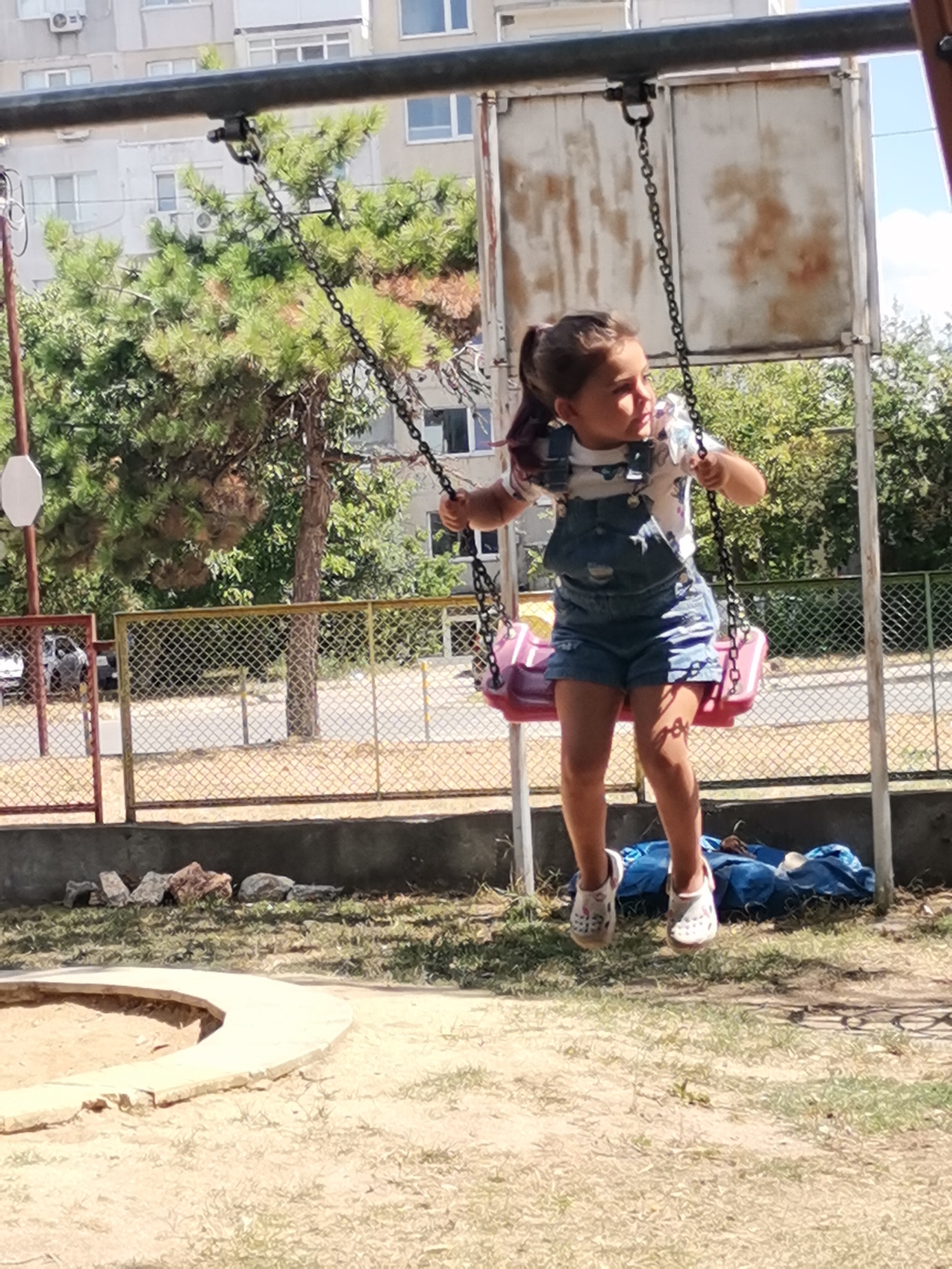 A young girl with brown hair pulled into a ponytail is swinging on a pink swing set. She is wearing denim overalls over a white t-shirt with a print. The swing set is attached to a metal frame, and the chains are visible. In the background, there is a building, trees, and a chain-link fence. The ground is covered in sand and some dry grass. The scene appears to be a playground in the city of Balchik, Bulgaria, indicated by the architecture of the building and the style of the playground equipment, common in Bulgarian urban areas.
