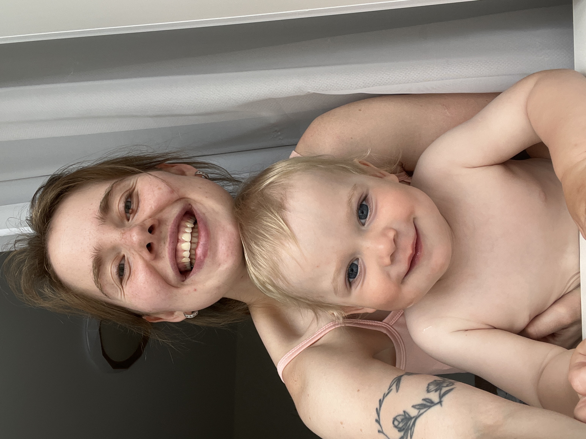 This image, captured in the city of Chaikovsky, Russia, shows a young woman holding a toddler in a close-up, indoor setting. The woman, positioned behind and slightly to the right, has light brown hair, light skin with visible freckles, and small silver hoop earrings. She is smiling broadly, exposing her upper teeth, and wears a light pink tank top. A tattoo depicting a floral wreath with roses and leaves is visible on her upper left arm. The toddler, with blonde hair and blue eyes, is in the foreground, smiling gently, and appears bare-chested. The woman's hands are visible supporting the child. The background on the left is dark, possibly a ceiling or upper wall, while the right side features a light grey curtain and a vertical white frame, indicating a room interior. Both individuals are looking directly towards the camera.