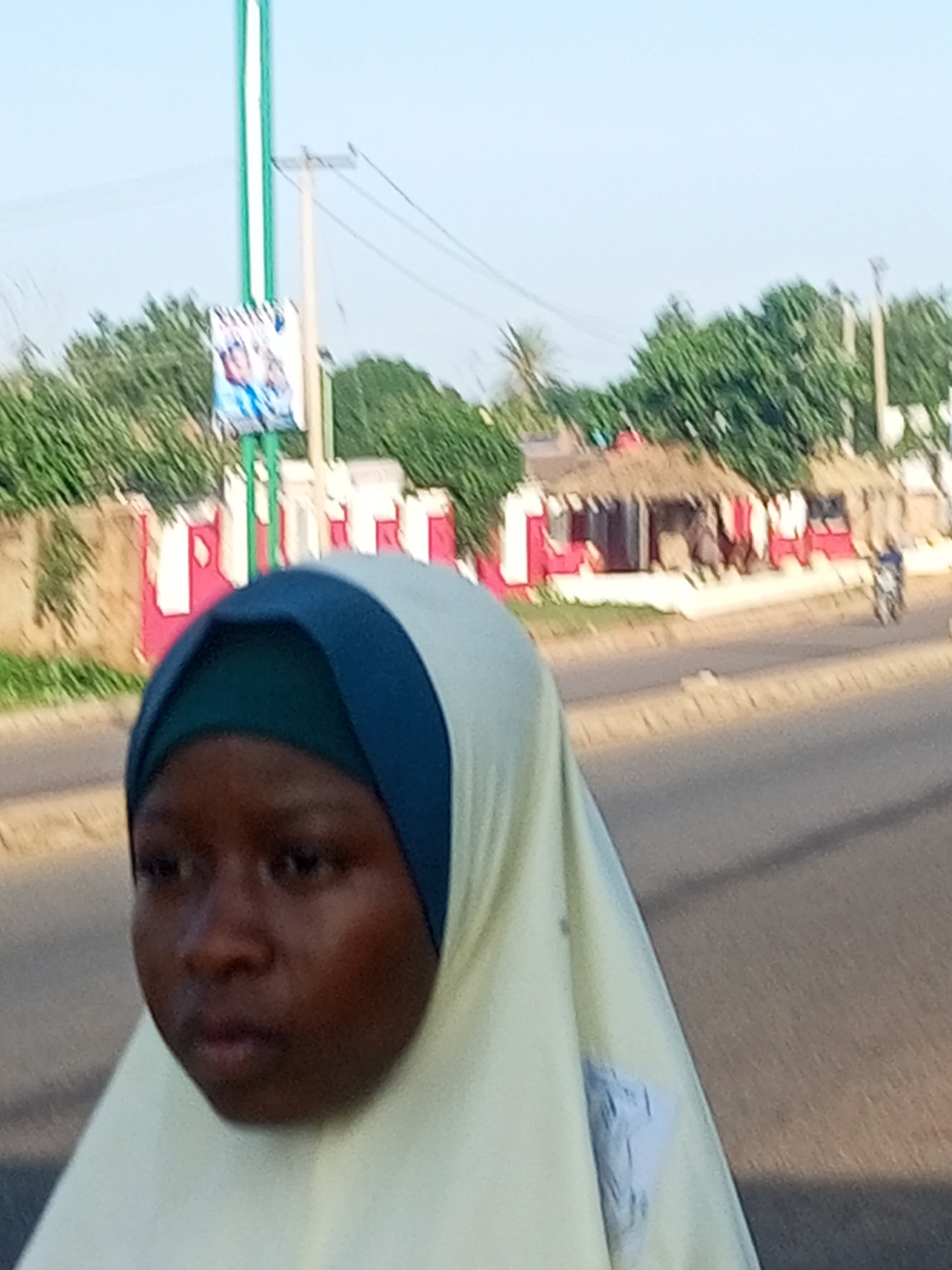 A young girl wearing a light yellow hijab with a dark blue band is the primary subject, positioned in the foreground. Her dark skin and prominent features are visible. She is facing to her left, looking slightly downwards. The background depicts a street scene with a paved road. Behind the road, there's a low wall, possibly a building facade adorned with red and white vertical stripes. Beyond that, a cluster of huts with thatched roofs is visible, with some laundry hanging outside. Lush green trees line the background, and utility poles with wires are also present. A light blue sky is above. To the right, a motorcycle is partially visible, suggesting movement in the scene. A green and white pole with a poster attached to it stands to the left of the girl, slightly out of focus. The overall impression is an outdoor, possibly informal urban or semi-urban environment in Tempure, Nigeria.