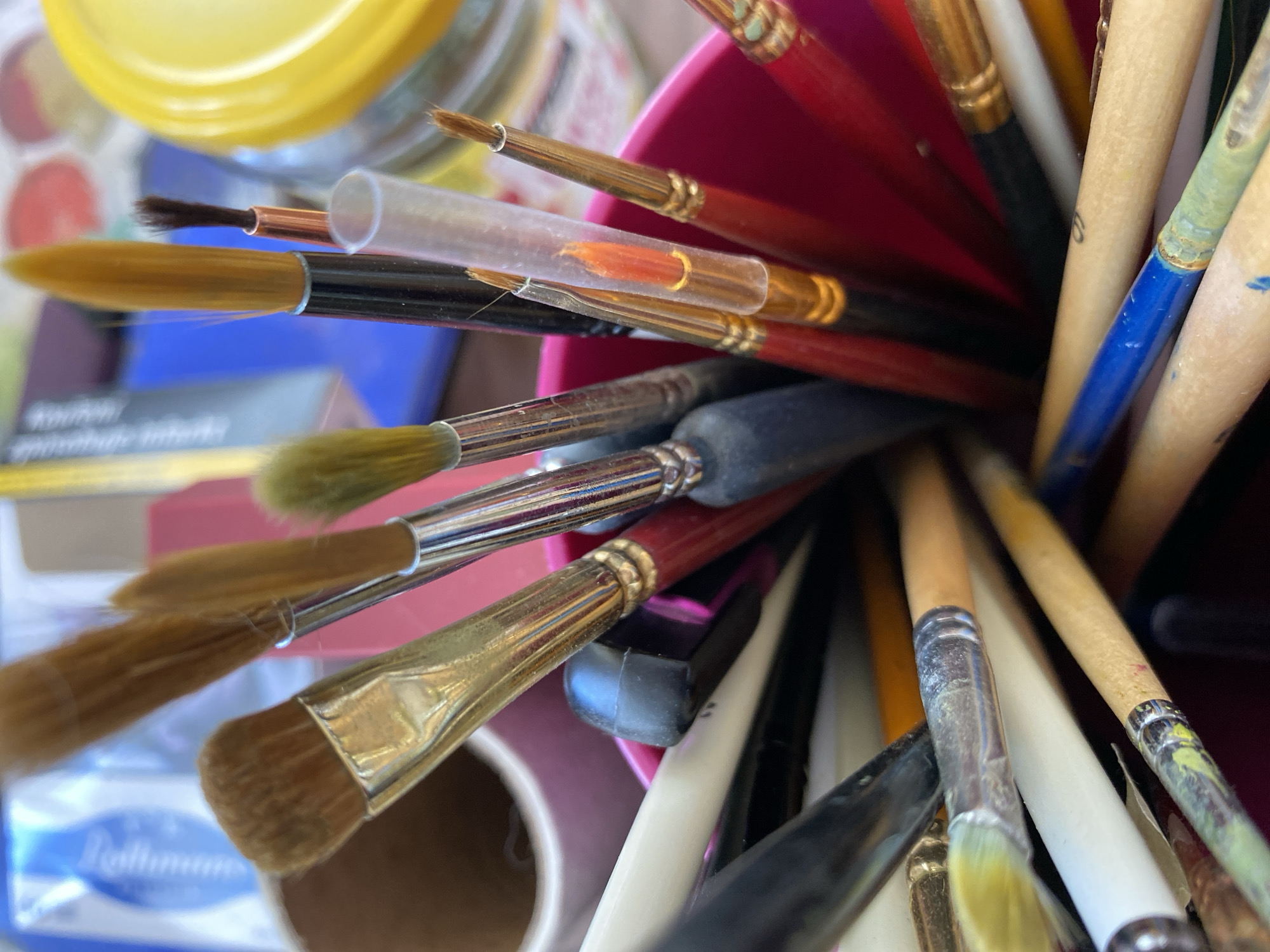 The image shows a close-up view of a collection of paintbrushes in a pink container. The brushes have various sizes and bristle types, with wooden and black handles. Some brushes have paint residue on them. In the background, out of focus, are art supplies such as tubes of paint and a yellow lid, possibly from a container of artist materials. The lighting suggests an indoor setting, possibly an art studio or a workspace. There are no people visible in the image. The image does not provide any information about the city of Bílina, Czechia.